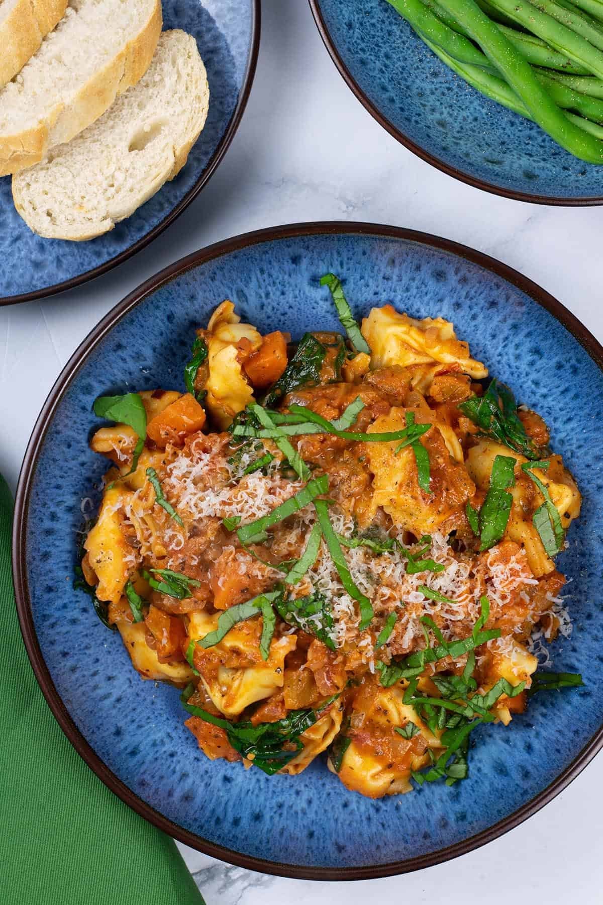 A portion of tortellini with creamy tomato and spinach sauce in a blue patterned bowl with a bowl of green beans and plate of sliced bread on the side.
