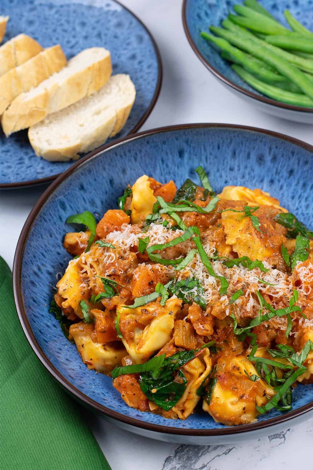 A portion of tortellini with creamy tomato and spinach sauce in a blue patterned bowl with a bowl of green beans and plate of sliced bread on the side.
