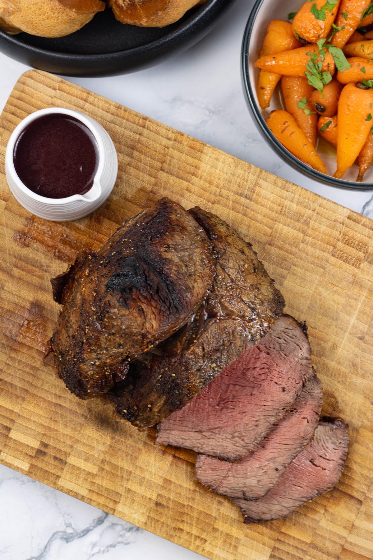 Air fryer roast beef sitting on a board with 3 slices carved, a small jug of red wine jus and bowls of Yorkshire puddings and chantenay carrots on the side.