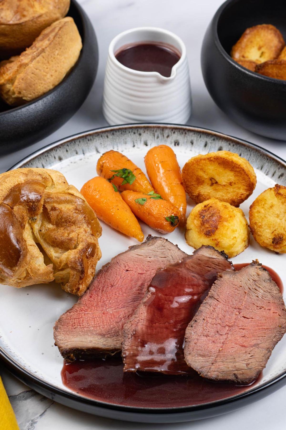 3 slices of air fryer roast beef on a black and white dinner plate with a Yorkshire pudding, chantenay carrots, roast potatoes and red wine jus.