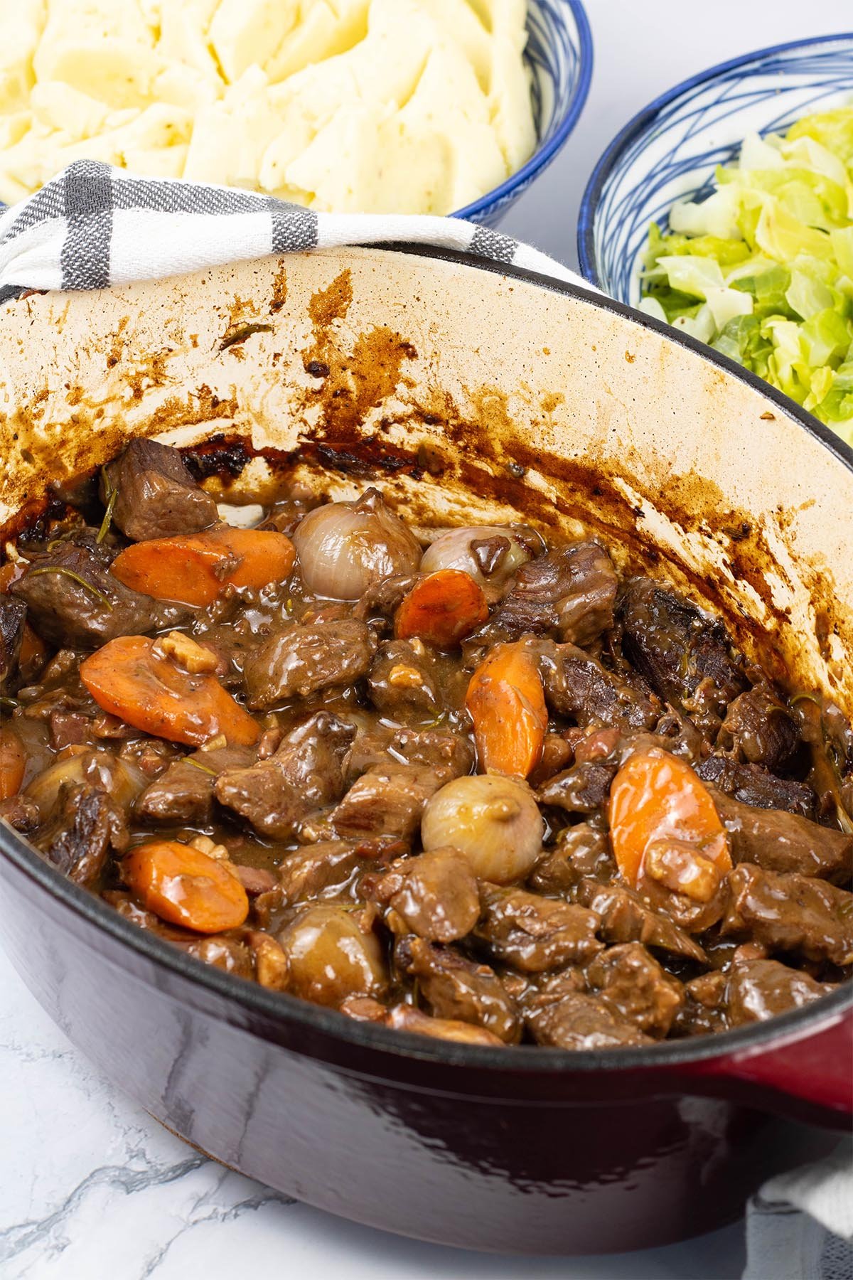 Beef bourguignon in a deep oval casserole dish with bowls of horseradish mash and buttered cabbage on the side.