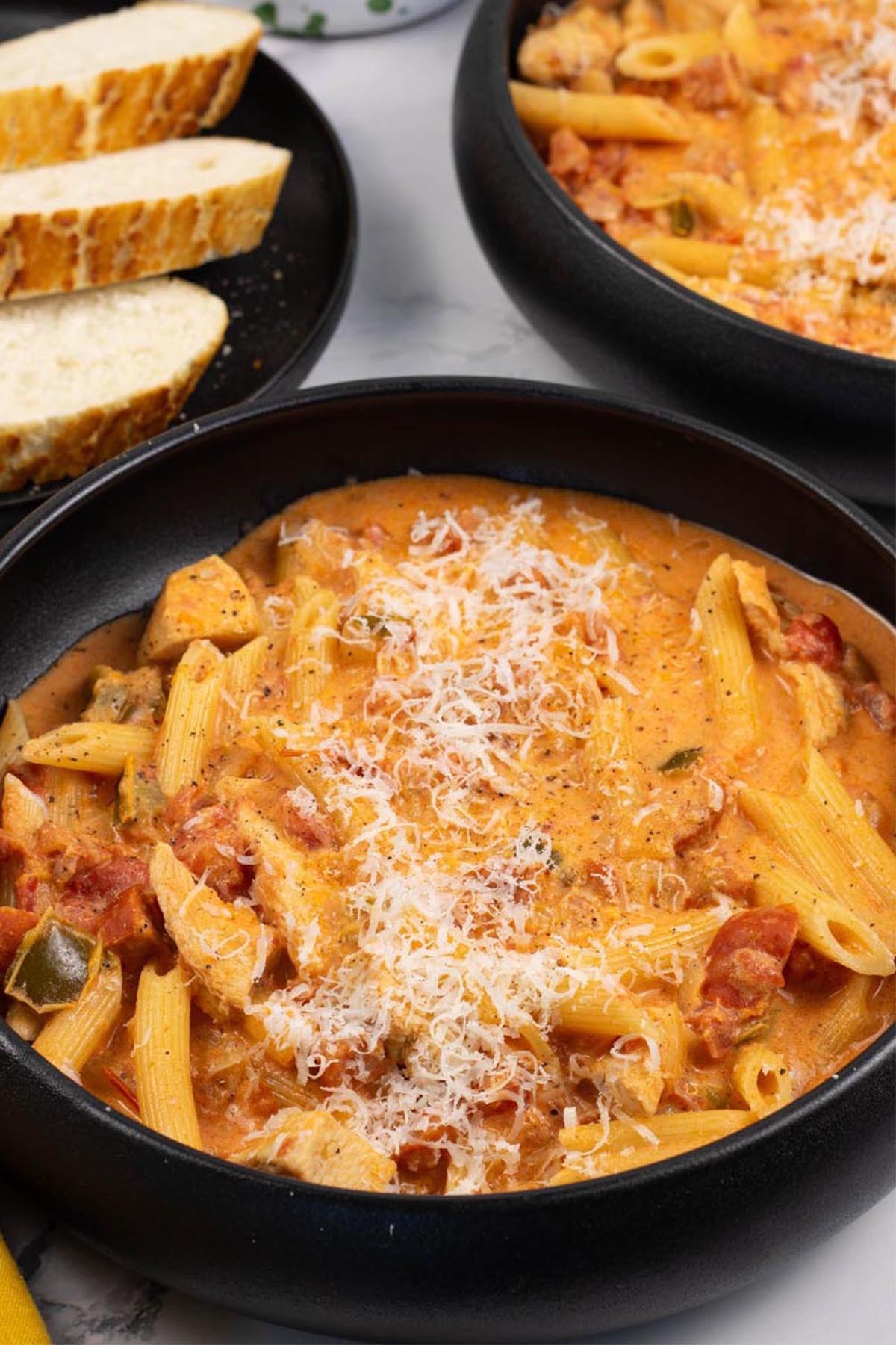 Two portions of cajun chicken and chorizo pasta in black bowls with a plate of sliced baguette on the side.