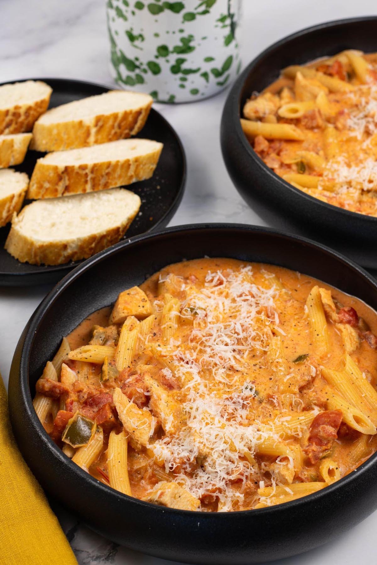 Two portions of cajun chicken and chorizo pasta in black bowls with a plate of sliced baguette on the side.