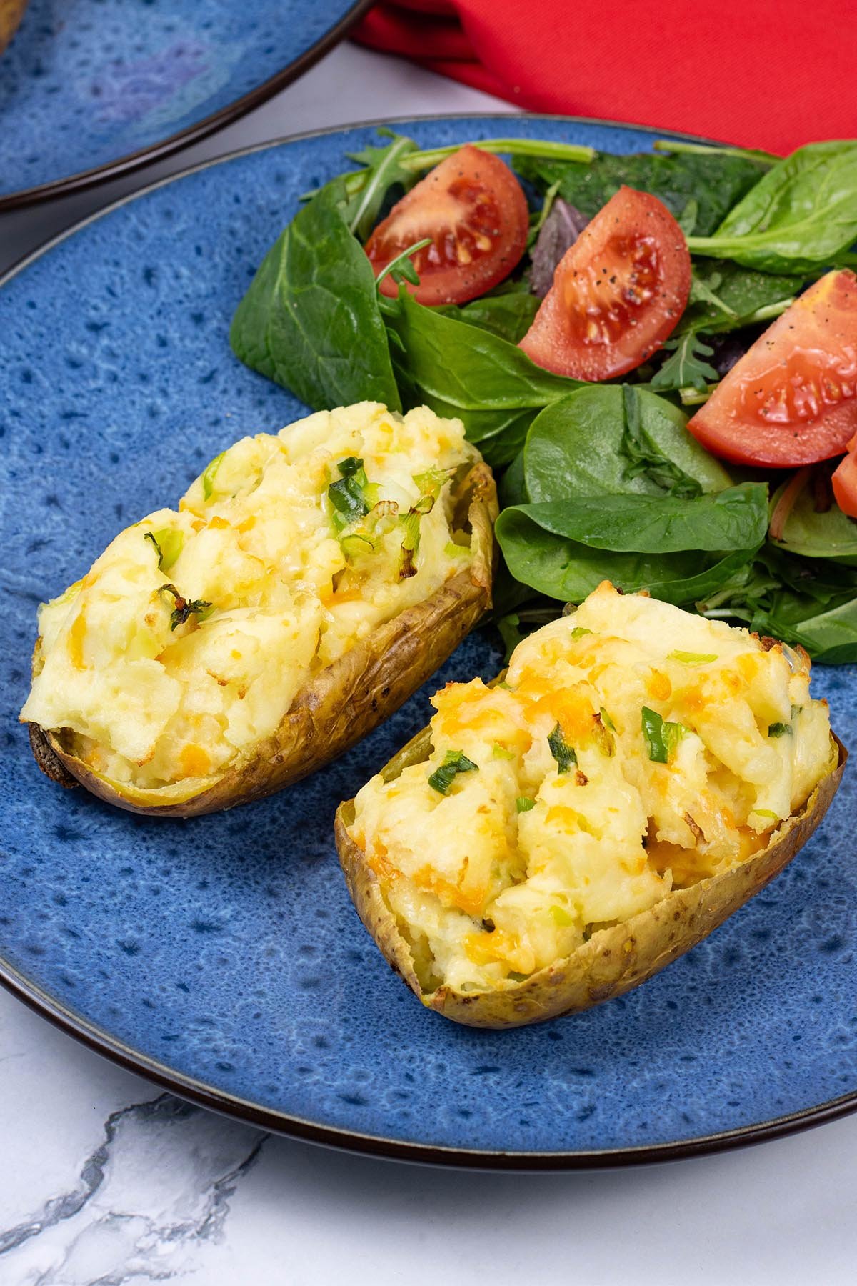 Cheese and spring onion baked potato on a blue patterned dinner plate with salad leaves and tomato wedges on the side.