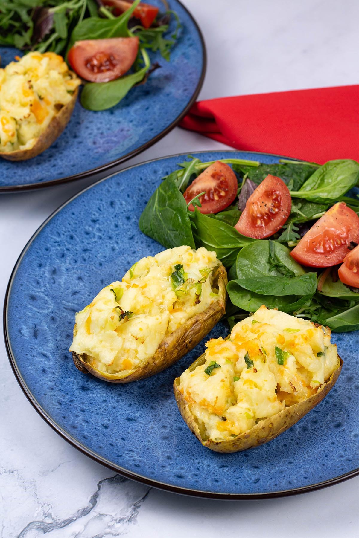 Cheese and spring onion baked potato on a blue patterned dinner plate with salad leaves and tomato wedges on the side.