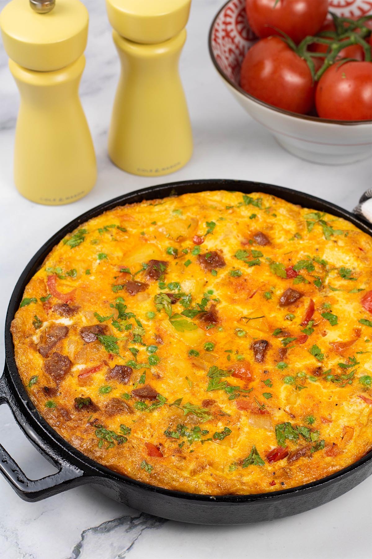Whole chorizo frittata in a medium frying pan with yellow salt and pepper grinders and bowl of tomatoes in the background.