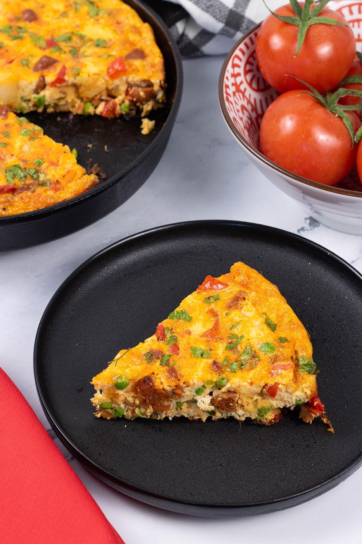 A slice of chorizo frittata on a black side plate with a red napkin on the side.