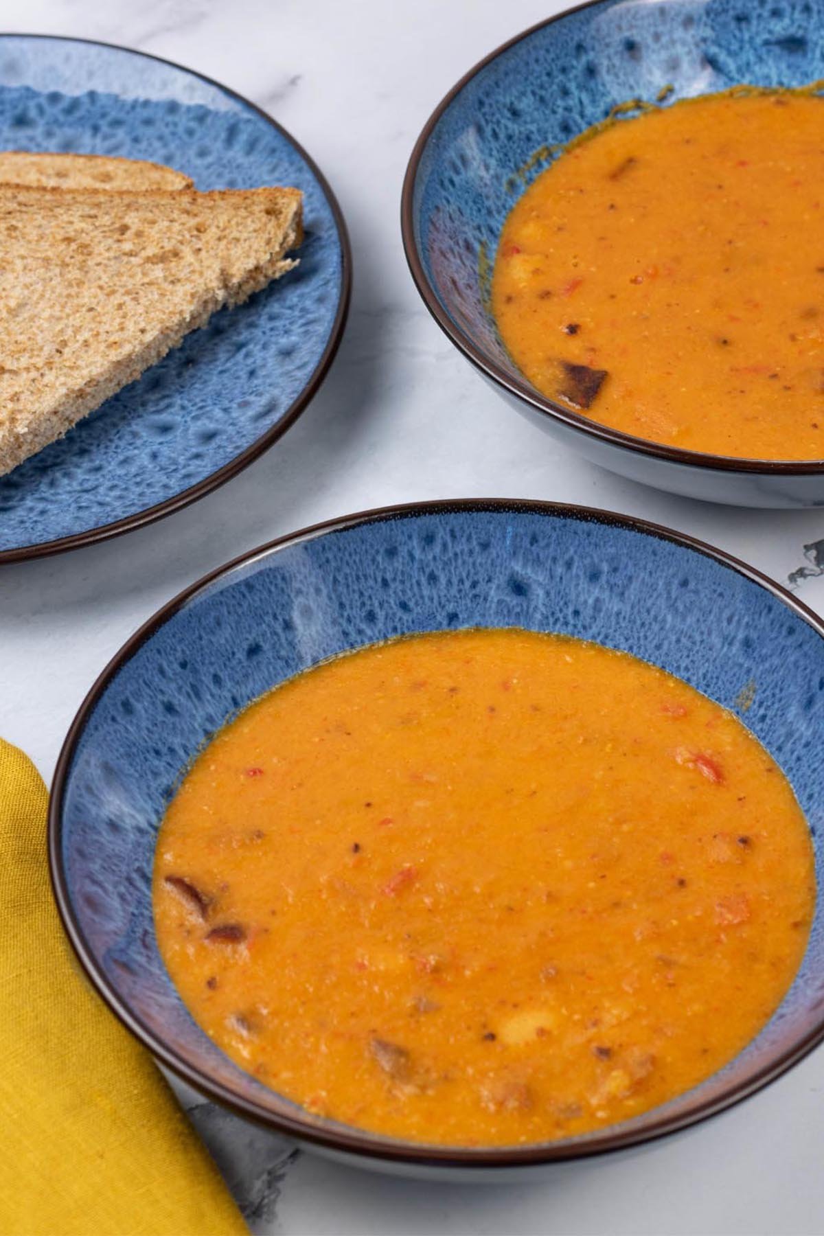 Two portions of chorizo and lentil soup in blue patterned bowls with a plate of sliced brown bread on the side.