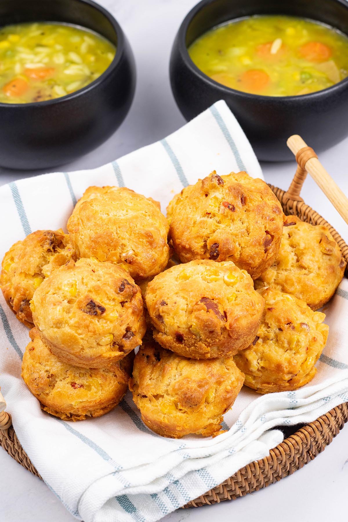 Chorizo, sweetcorn and chilli muffins in a bread basket, with 2 bowls of chunky vegetable soup in the background.