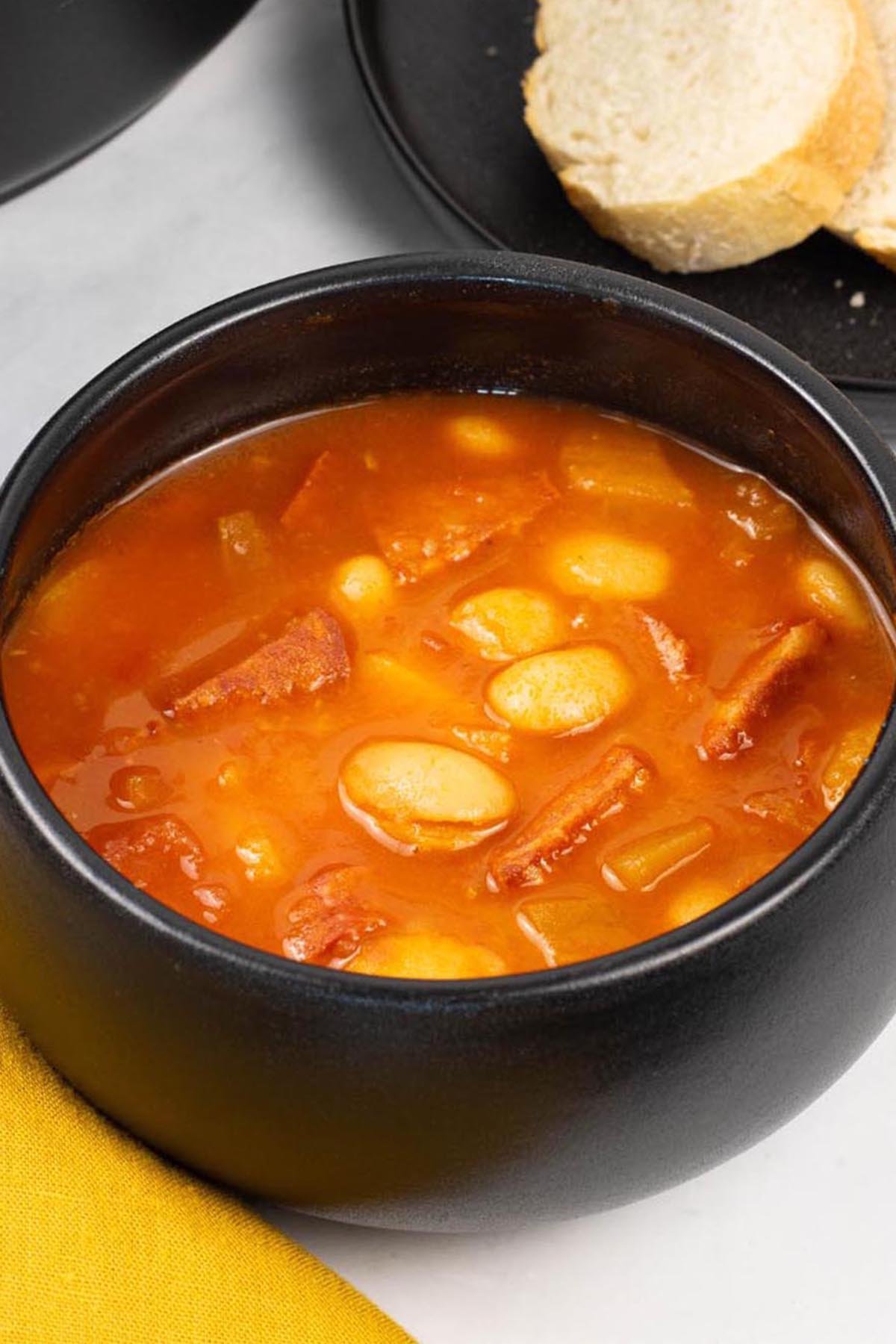 A portion of chunky chorizo and butter bean soup in a black bowl with a plate of sliced crusty bread on the side.