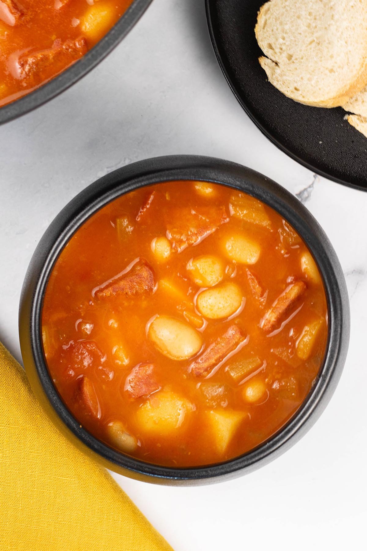 A portion of chunky chorizo and butter bean soup in a black bowl with a plate of sliced crusty bread on the side.