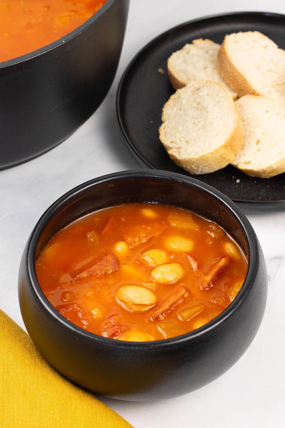 A portion of chunky chorizo and butter bean soup in a black bowl with a plate of sliced crusty bread on the side.
