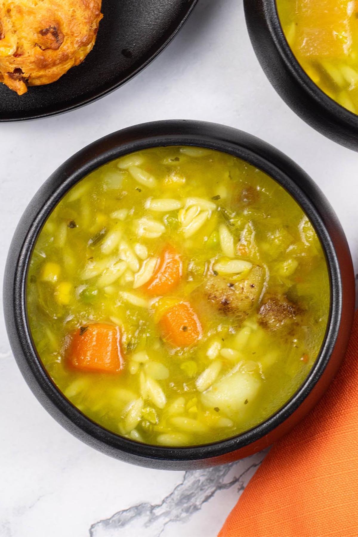 Two portions of chunky mixed vegetable soup made in the Thermomix, in black bowls with a plate of chorizo, sweetcorn and chilli muffins on the side.