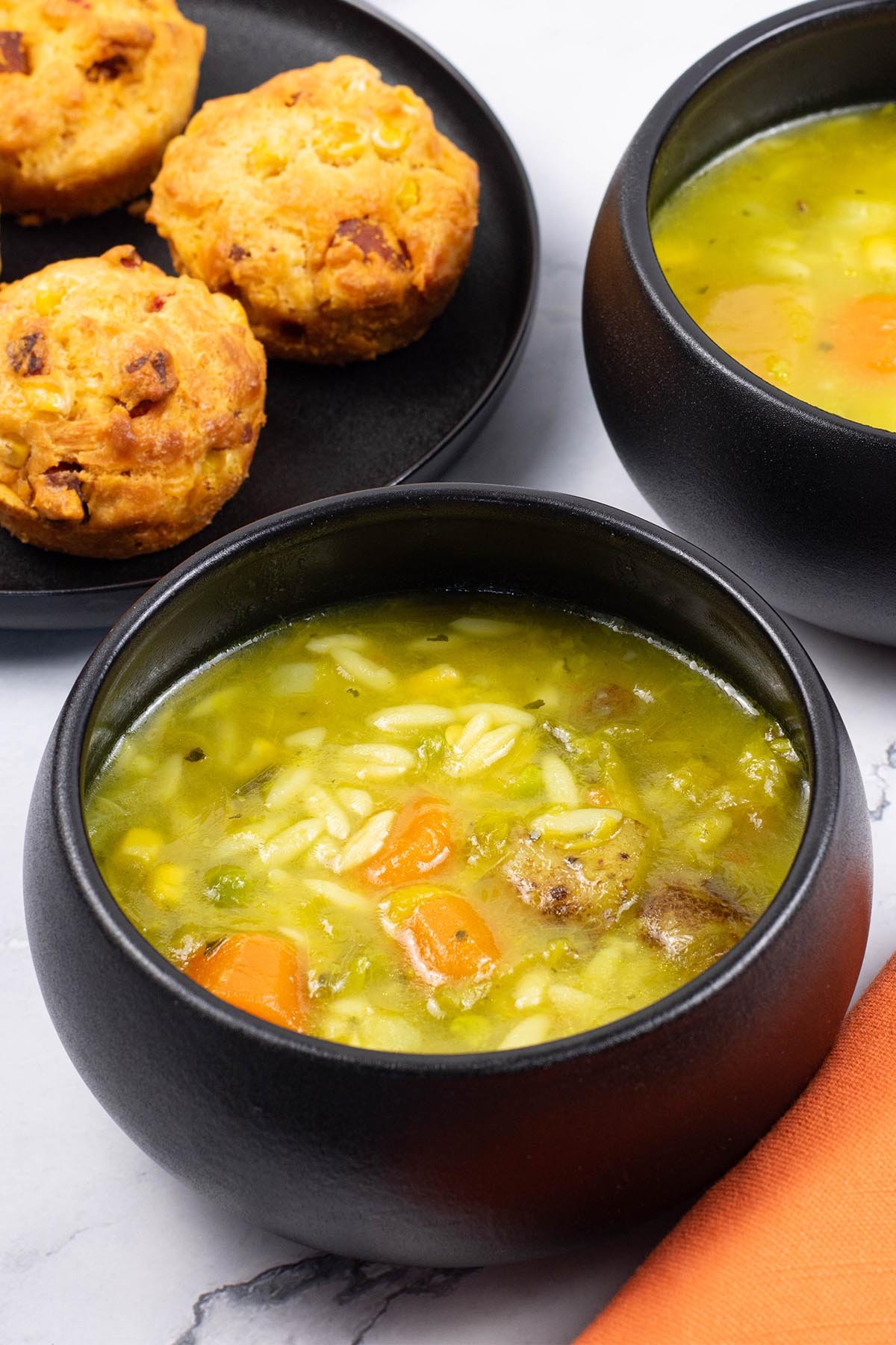 Two portions of chunky mixed vegetable soup made in the Thermomix, in black bowls with a plate of chorizo, sweetcorn and chilli muffins on the side.