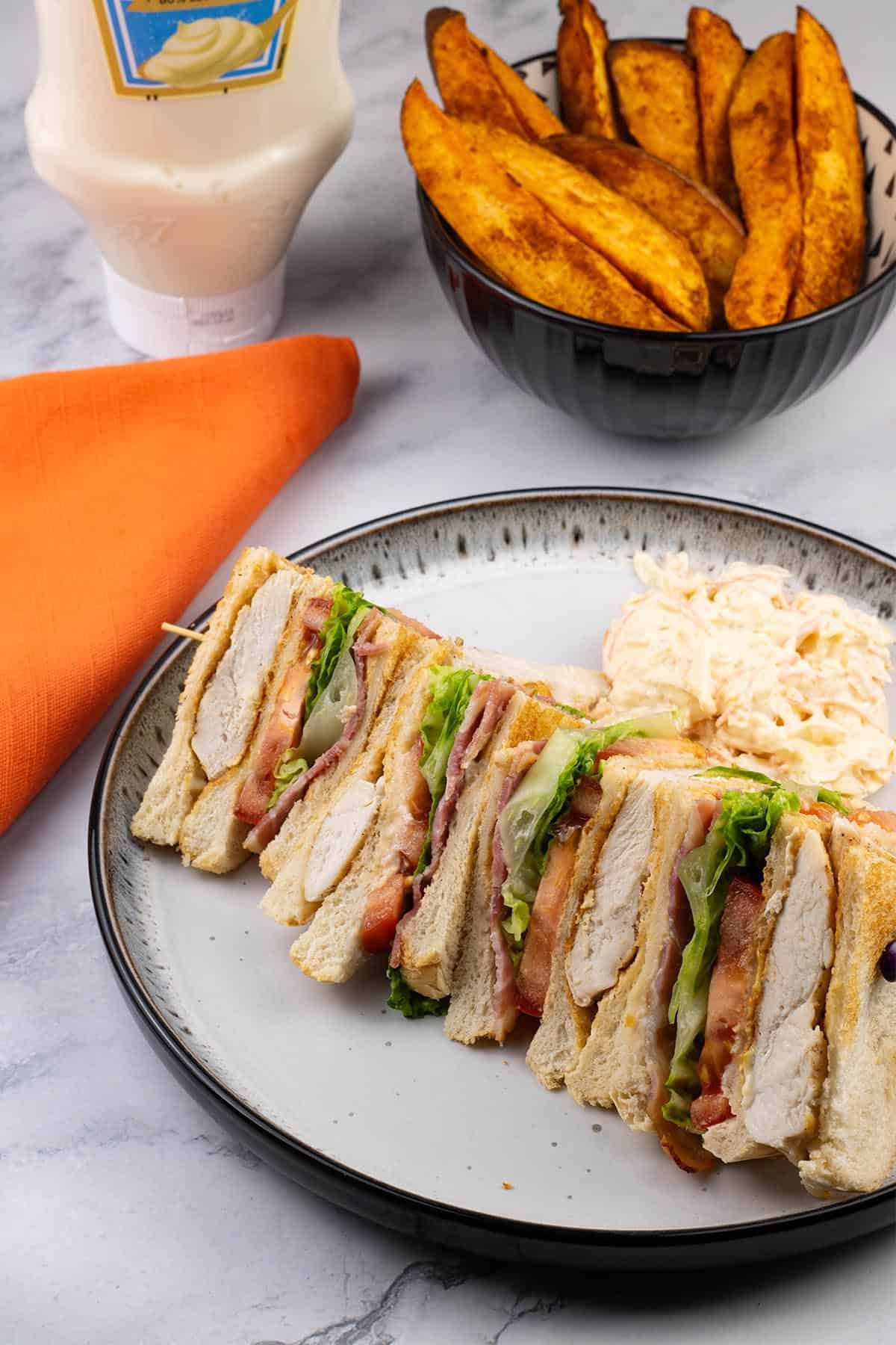 A club sandwich and coleslaw on a black and white patterned dinner plate with a bowl of sweet potato wedges, bottle of mayo and orange napkin on the side.