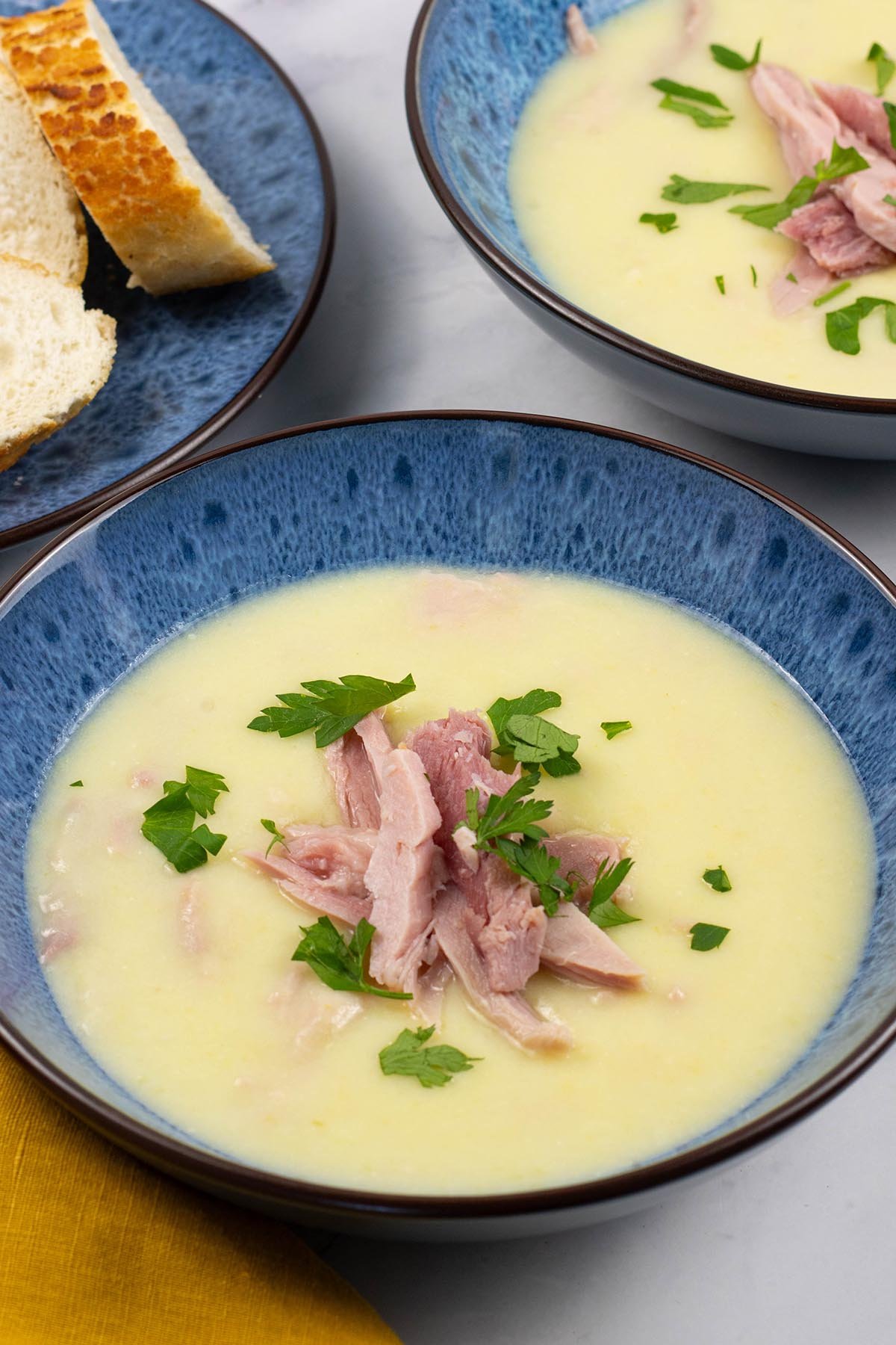 Two portions of creamy leek and potato soup with ham in blue patterned bowls, with a plate of sliced baguette on the side.