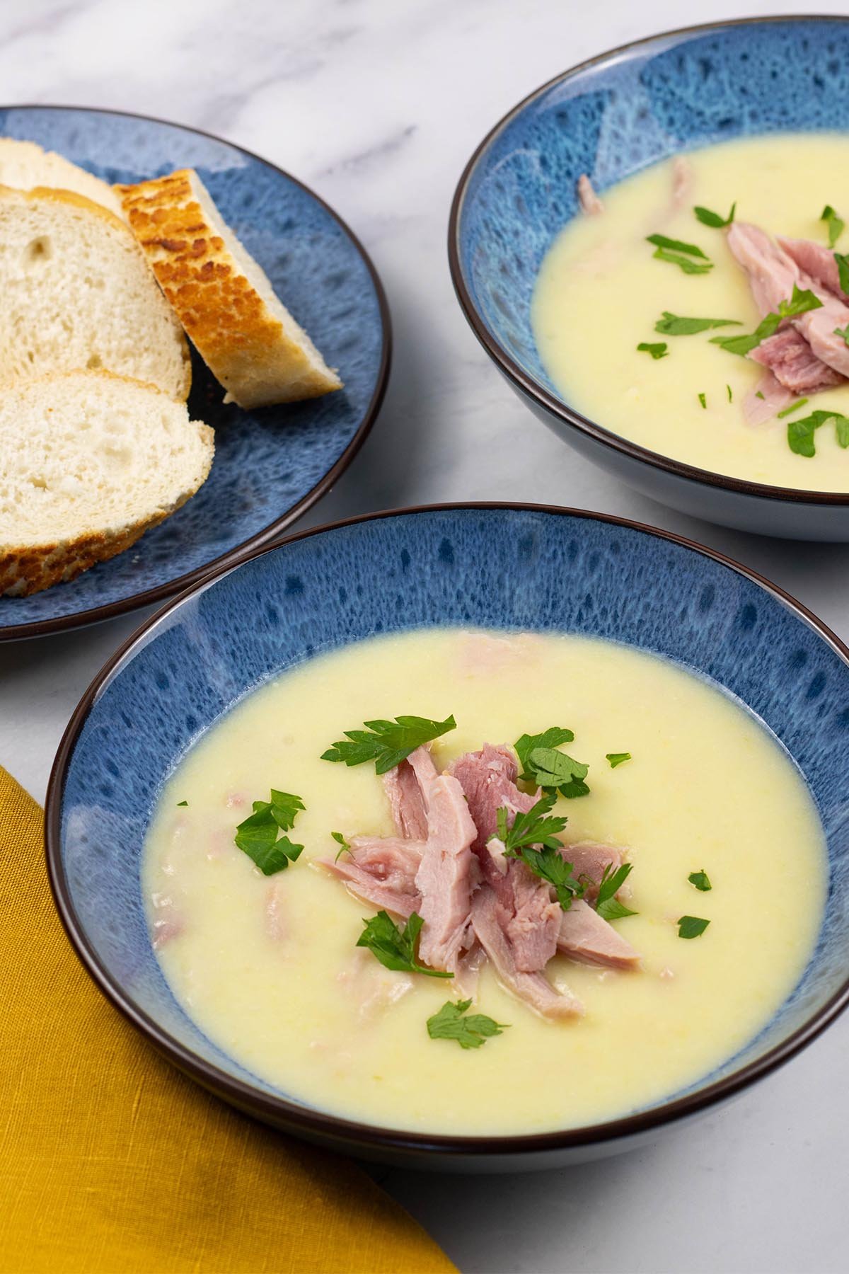 Two portions of creamy leek and potato soup with ham in blue patterned bowls, with a plate of sliced baguette on the side.