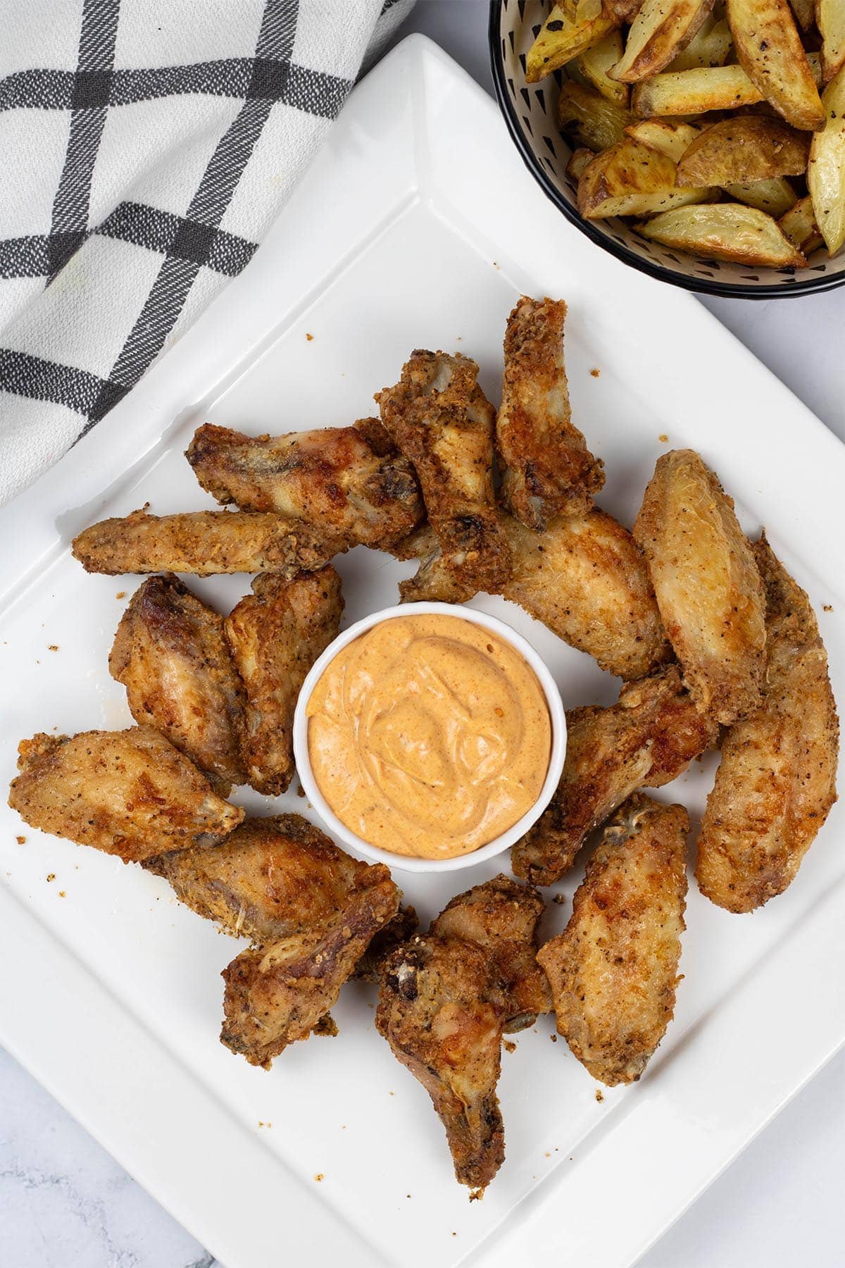 Easy air fryer cajun chicken wings on a white square platter with a dish of cajun dipping sauce and a bowl of chunky oven chips on the side.