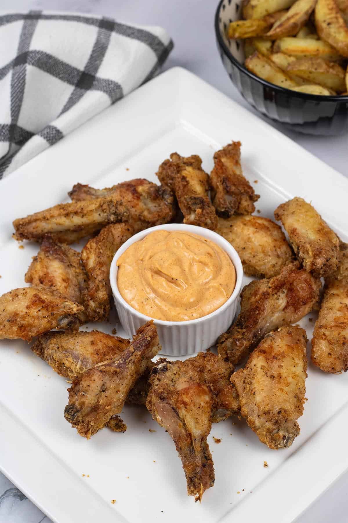 Easy air fryer cajun chicken wings on a white square platter with a dish of cajun dipping sauce and a bowl of chunky oven chips on the side.