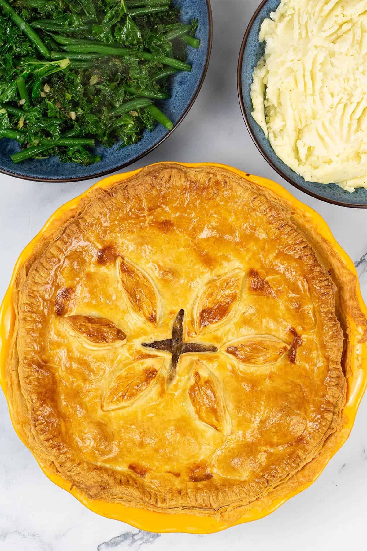 Easy steak pie in a round yellow pie dish with bowls of mashed potatoes, and sautéed kale and green beans on the side.