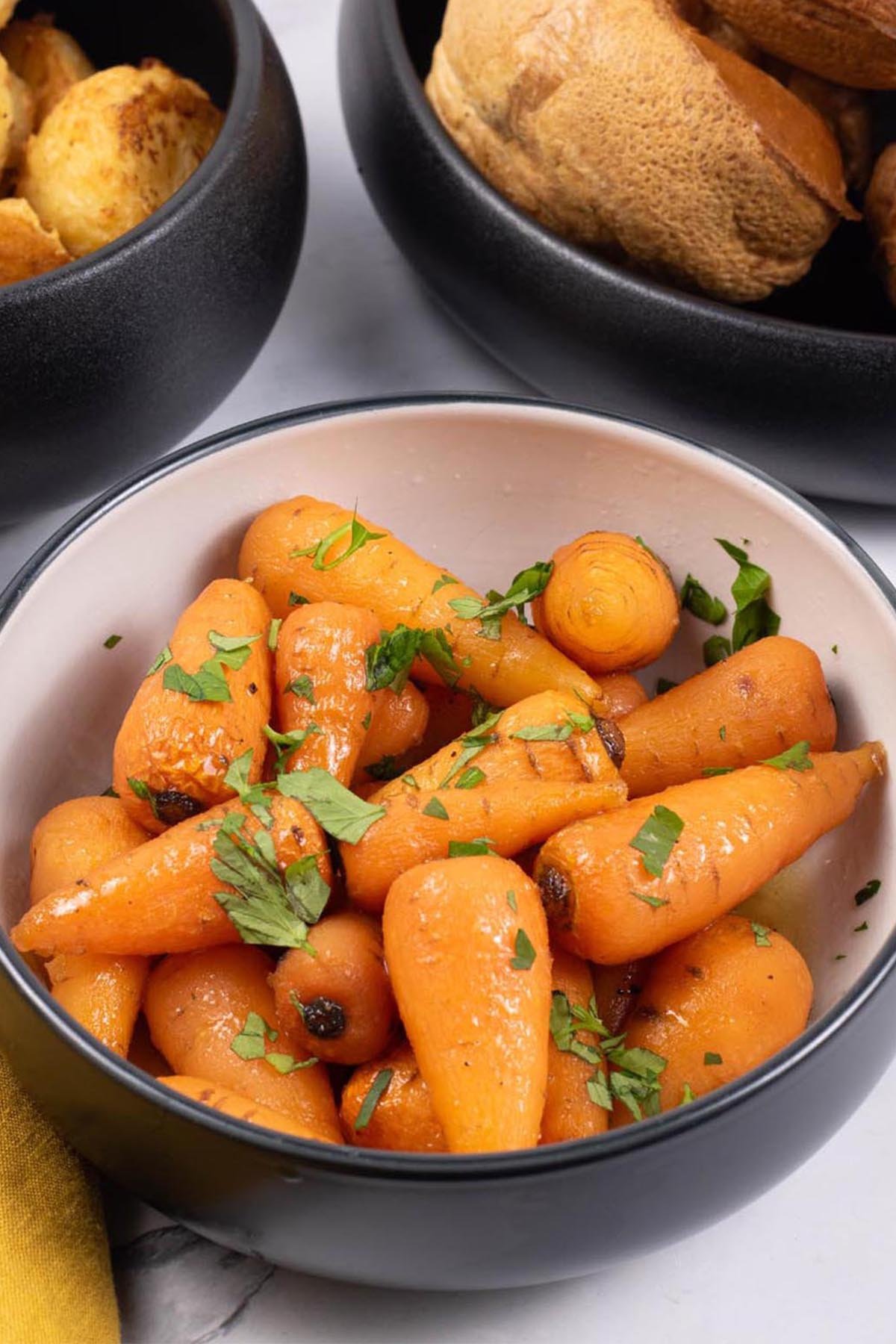 Easy sweet roasted chantenay carrots in a black and white serving bowl, with bowls of roast potatoes and Yorkshire puddings on the side.