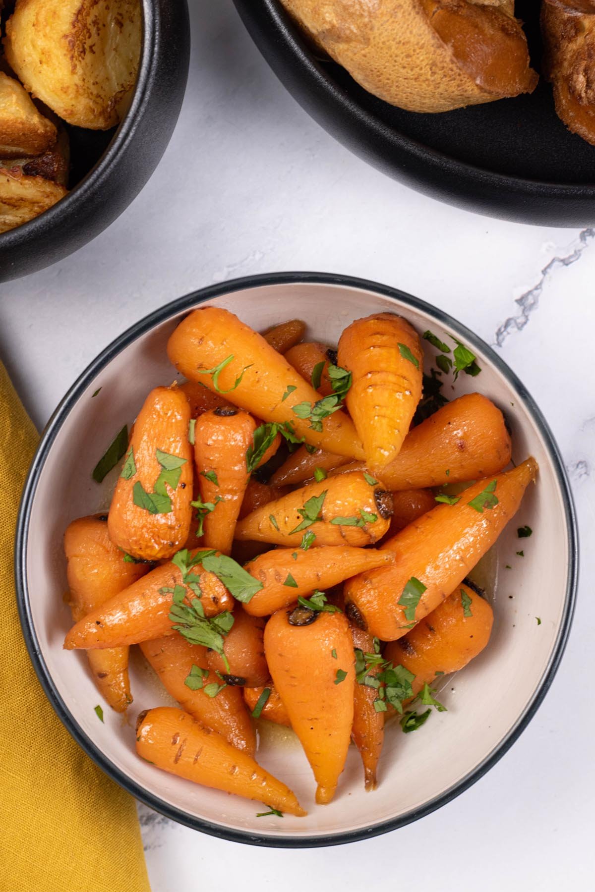Easy sweet roasted chantenay carrots in a black and white serving bowl, with bowls of roast potatoes and Yorkshire puddings on the side.