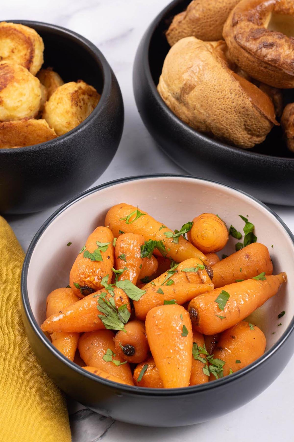 Easy sweet roasted chantenay carrots in a black and white serving bowl, with bowls of roast potatoes and Yorkshire puddings on the side.