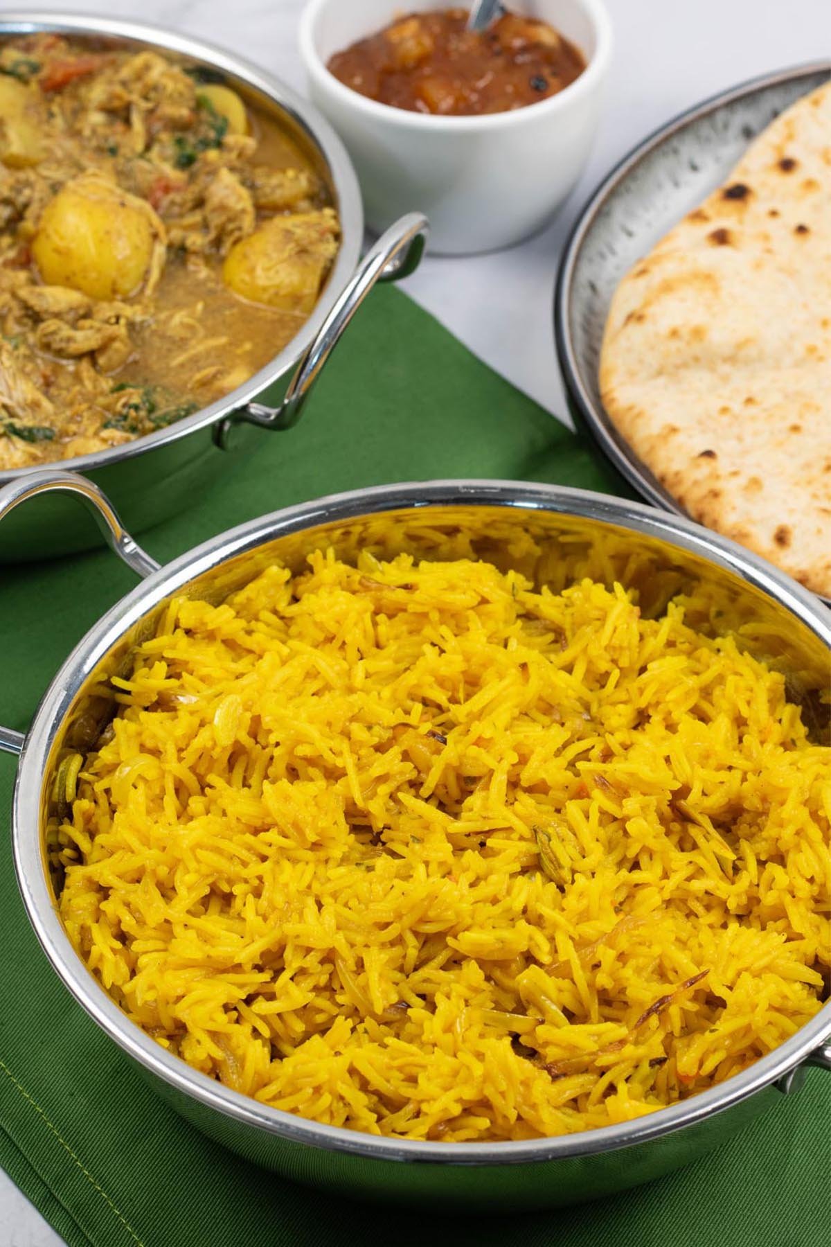 Golden pilau rice in a balti dish with a dish of slow cooker chicken and potato curry, bowl of mango chutney and plate of chapatis in the background.