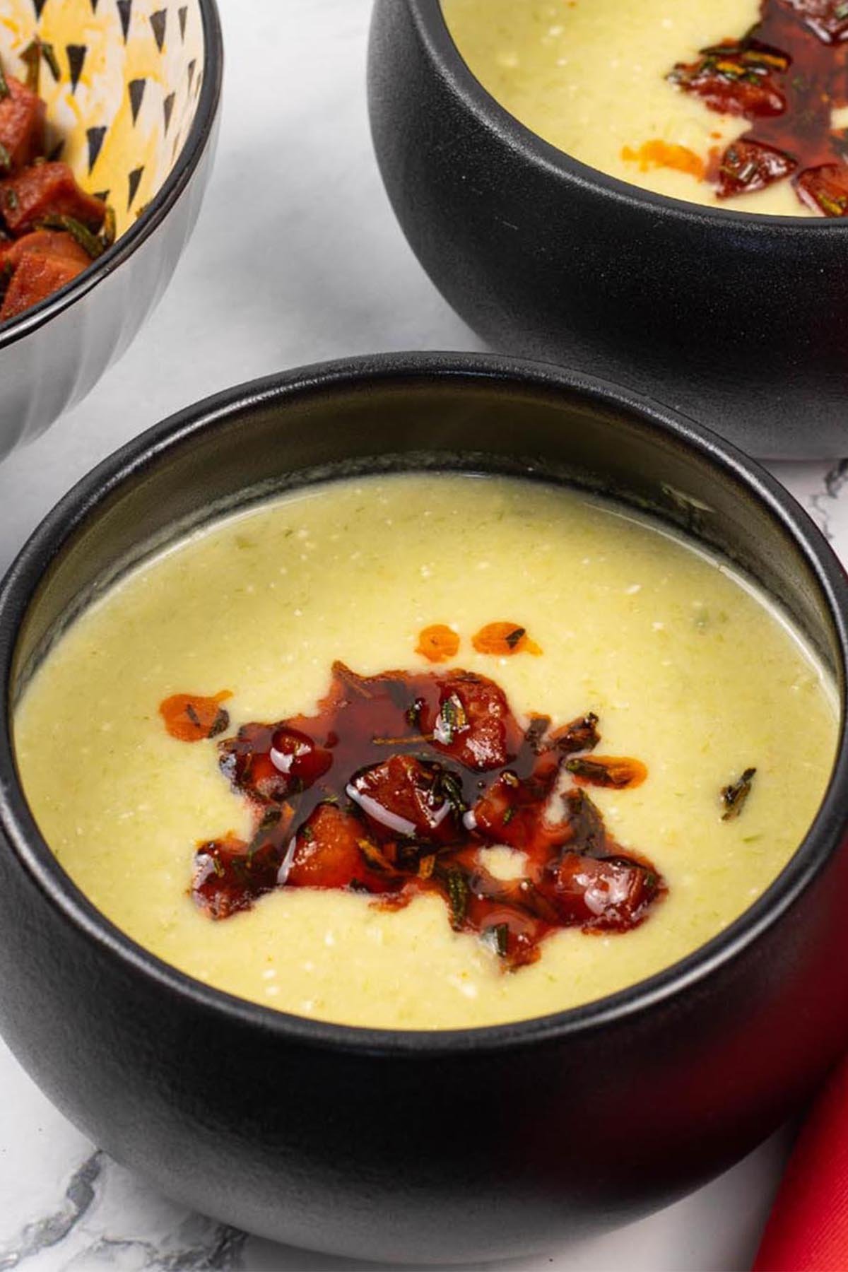 Two portions of leek, butter bean and crispy chorizo soup in black bowls with a bowl of crispy chorizo on the side.