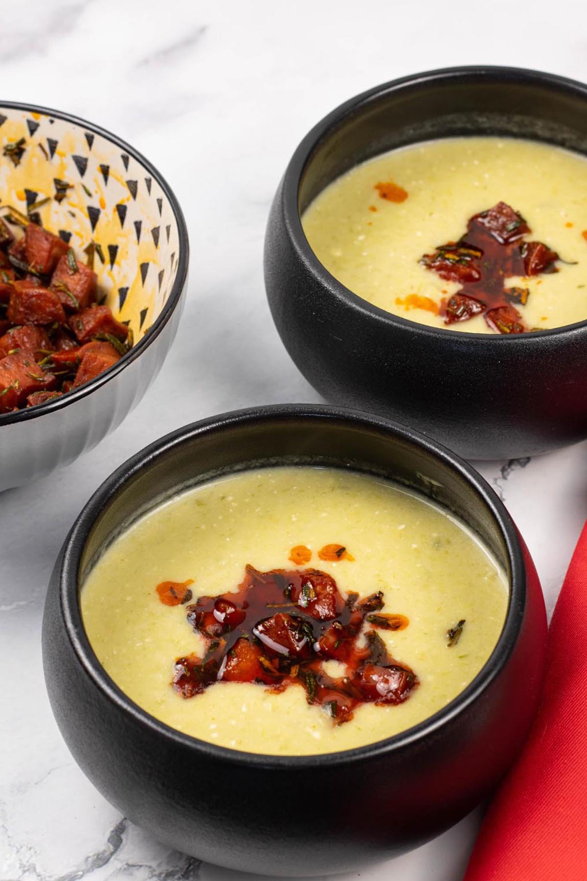 Two portions of leek, butter bean and crispy chorizo soup in black bowls with a bowl of crispy chorizo on the side.