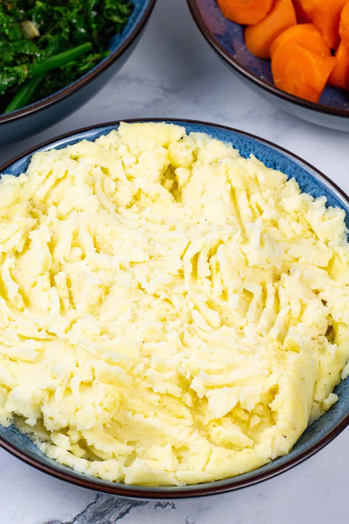 Mashed potatoes in a blue patterned serving bowl with bowls of sautéed kale and green beans, and quick cook carrots in the background.
