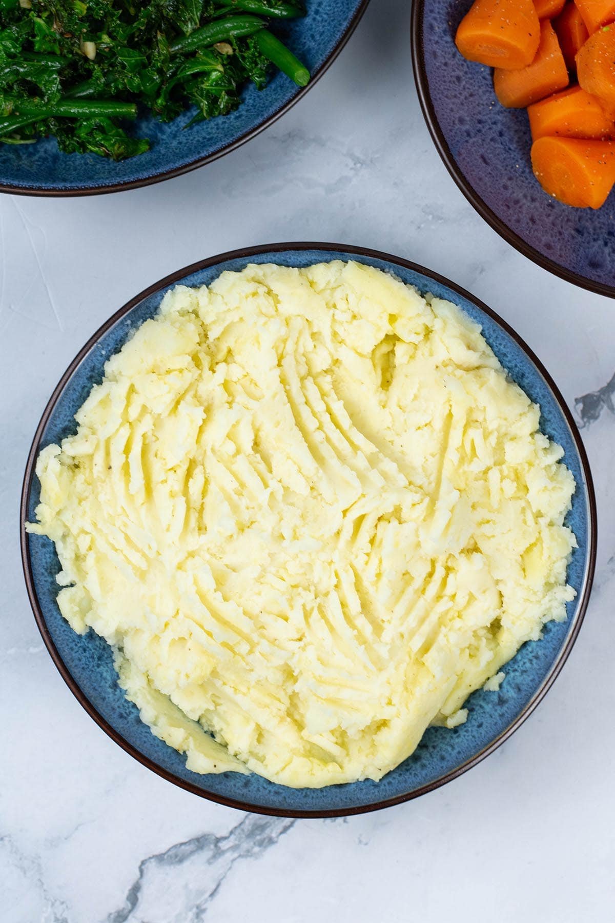 Mashed potatoes in a blue patterned serving bowl with bowls of sautéed kale and green beans, and quick cook carrots in the background.