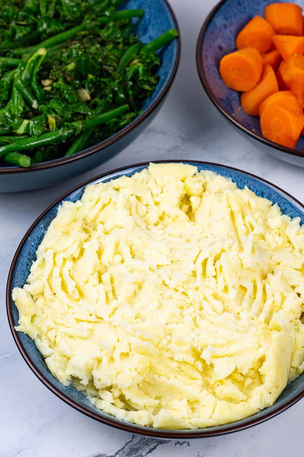Mashed potatoes in a blue patterned serving bowl with bowls of sautéed kale and green beans, and quick cook carrots in the background.