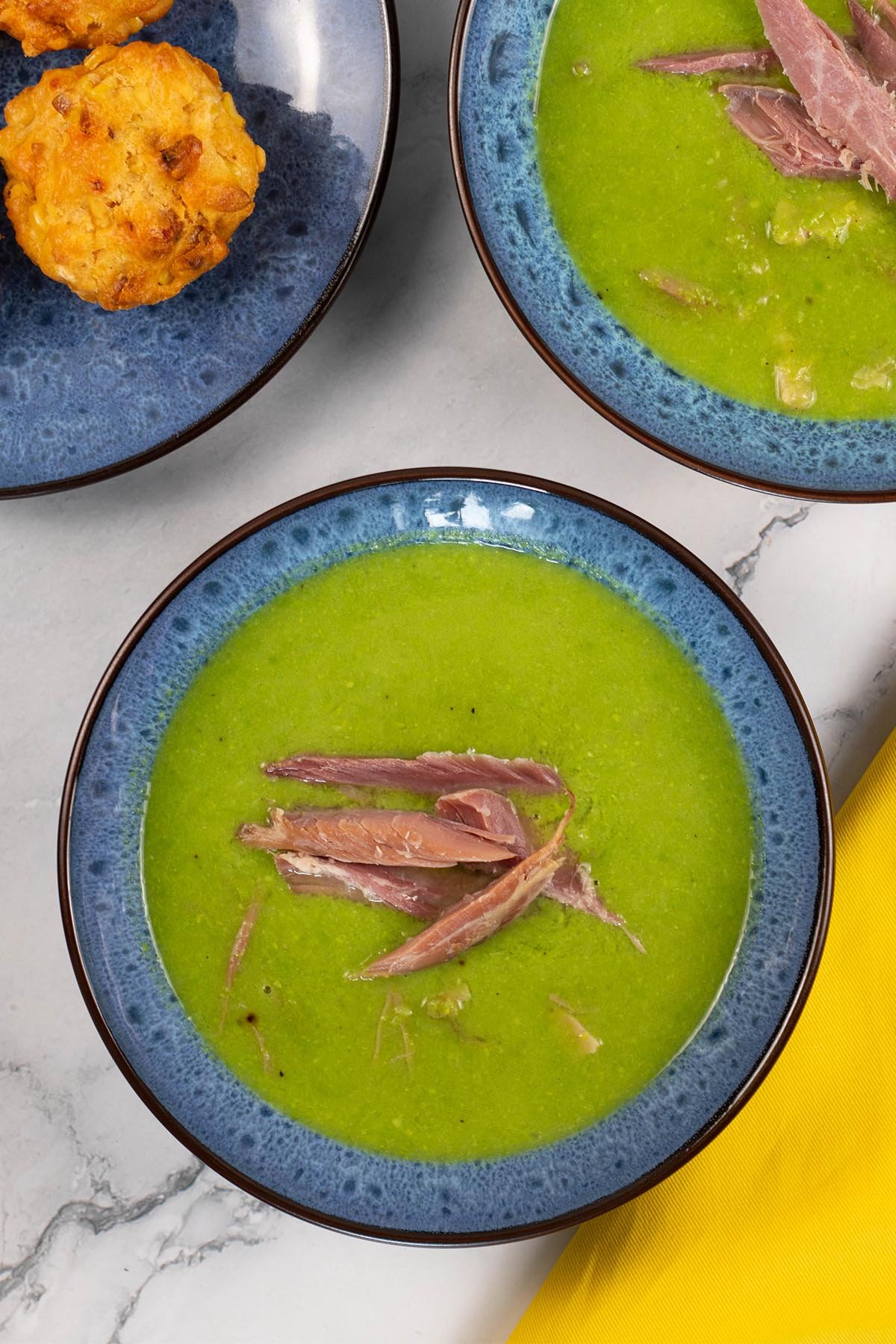Two portions of pea and ham soup in blue patterned bowls with a plate of chorizo, sweetcorn and chilli muffins on the side.