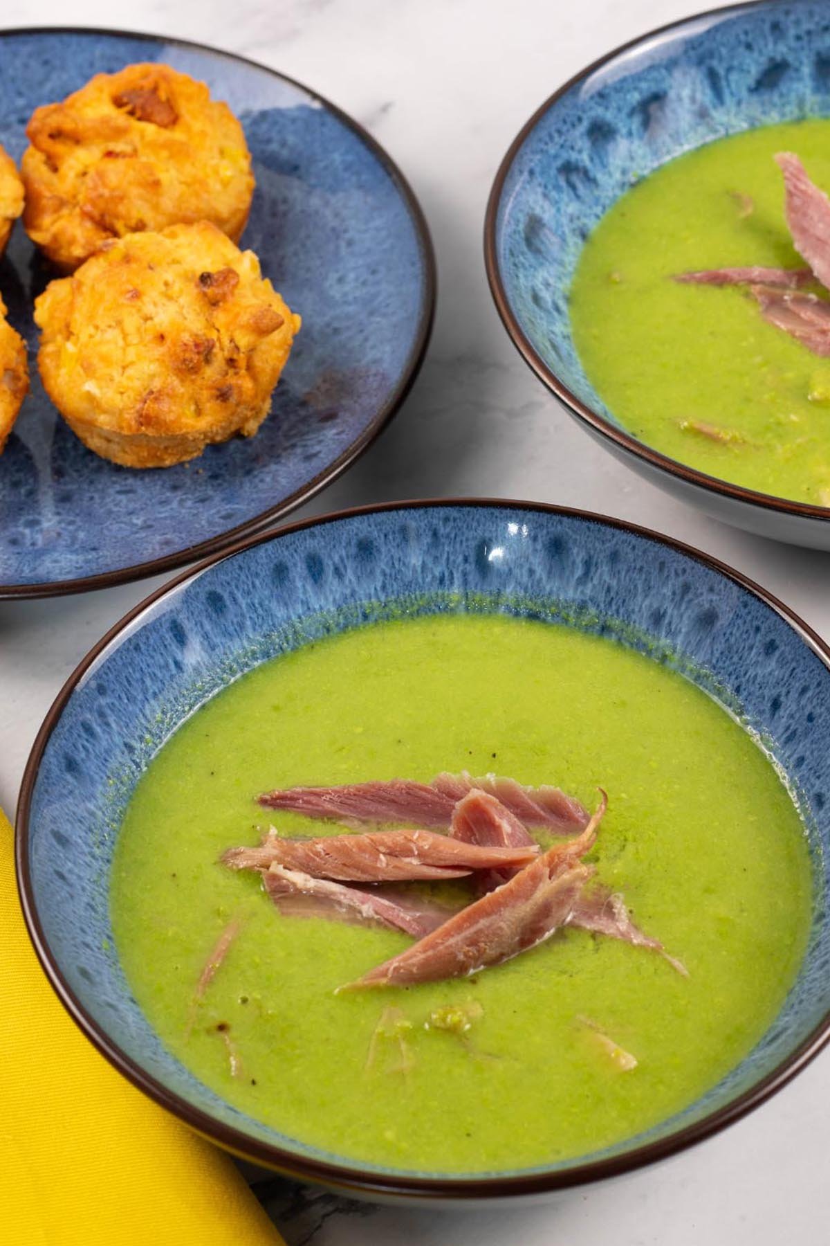 Two portions of pea and ham soup in blue patterned bowls with a plate of chorizo, sweetcorn and chilli muffins on the side.