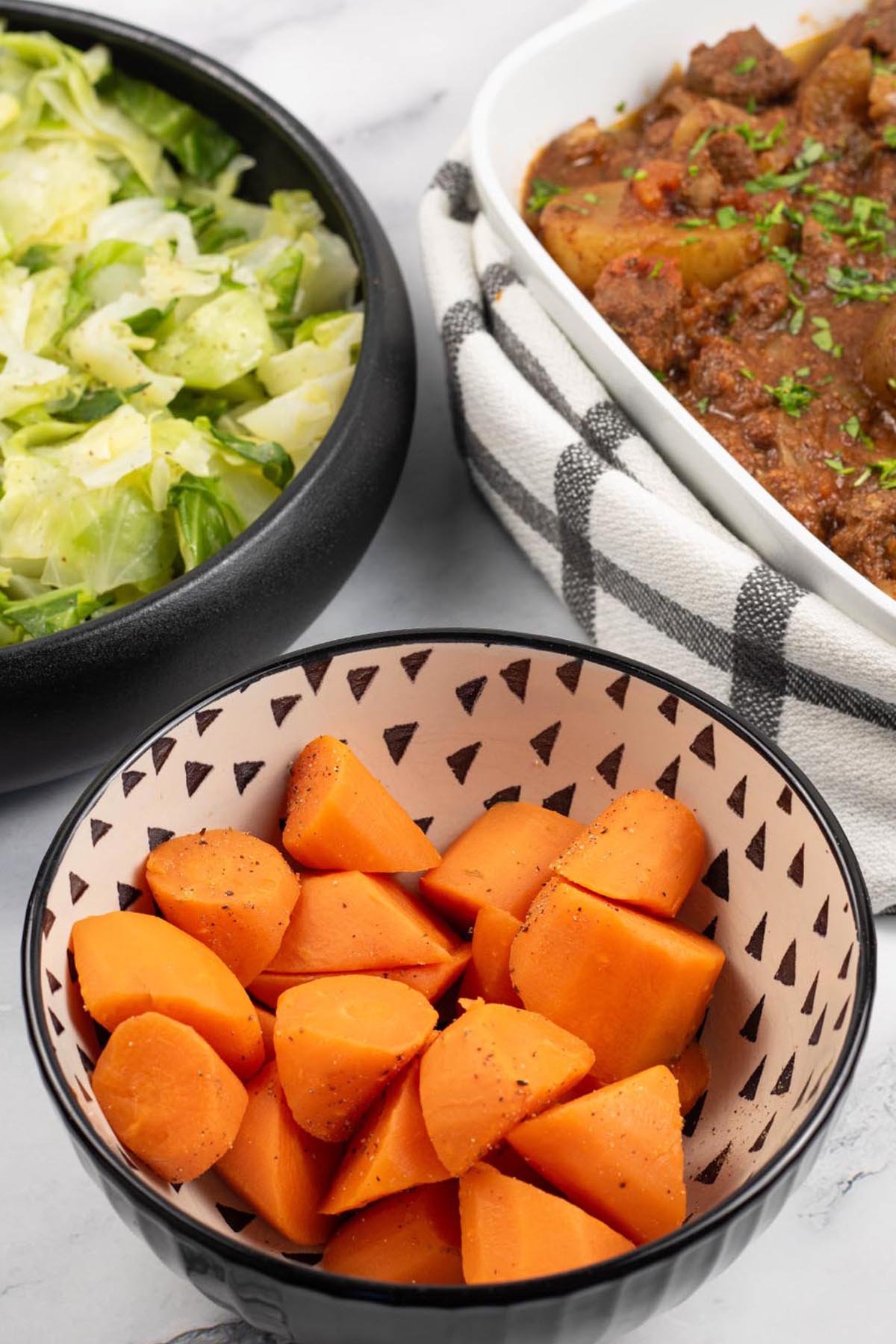 Quick cook carrots in a black and cream patterned serving bowl with dishes of buttered cabbage and slow cooker lamb stew in the background.