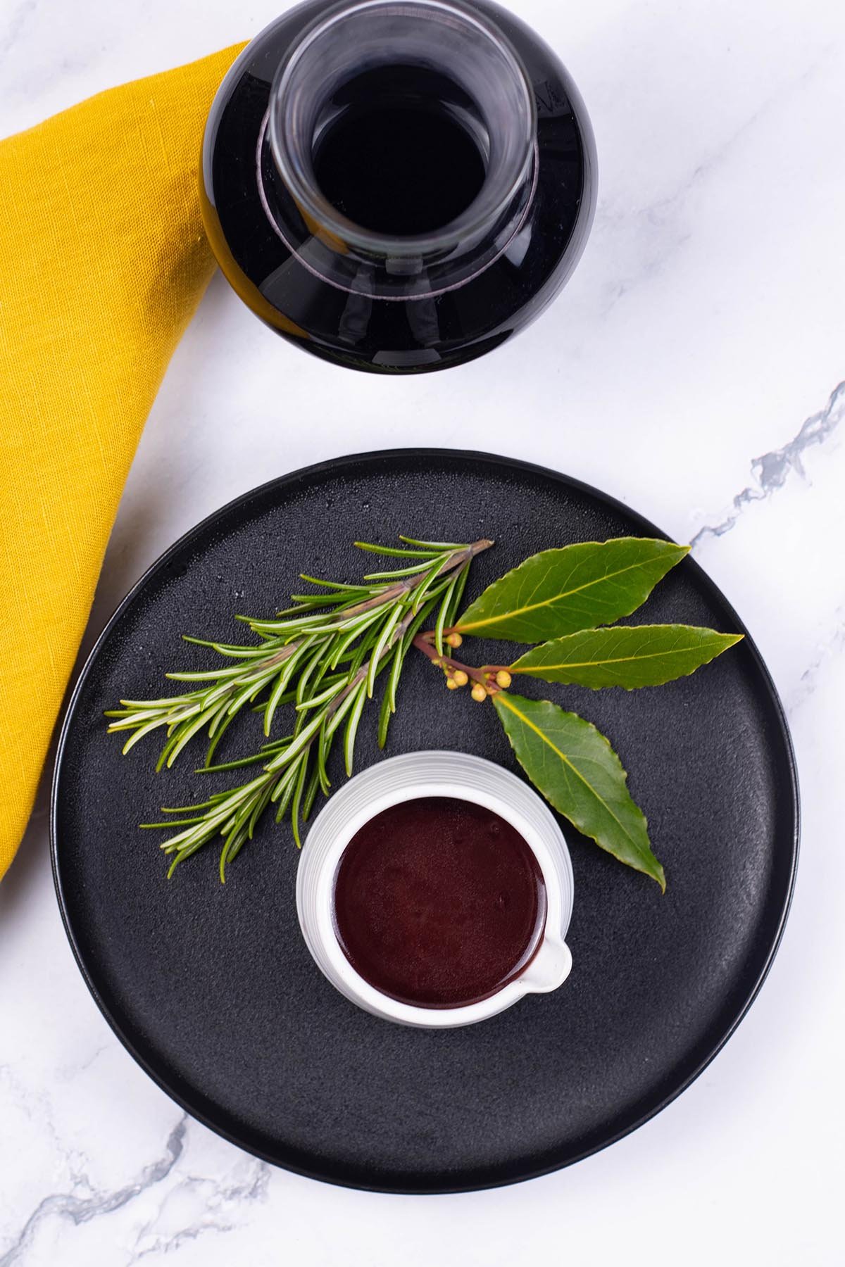Red wine jus in a small white ribbed jug sitting on a black side plate decorated with 2 sprigs of rosemary and 3 bay leaves.
