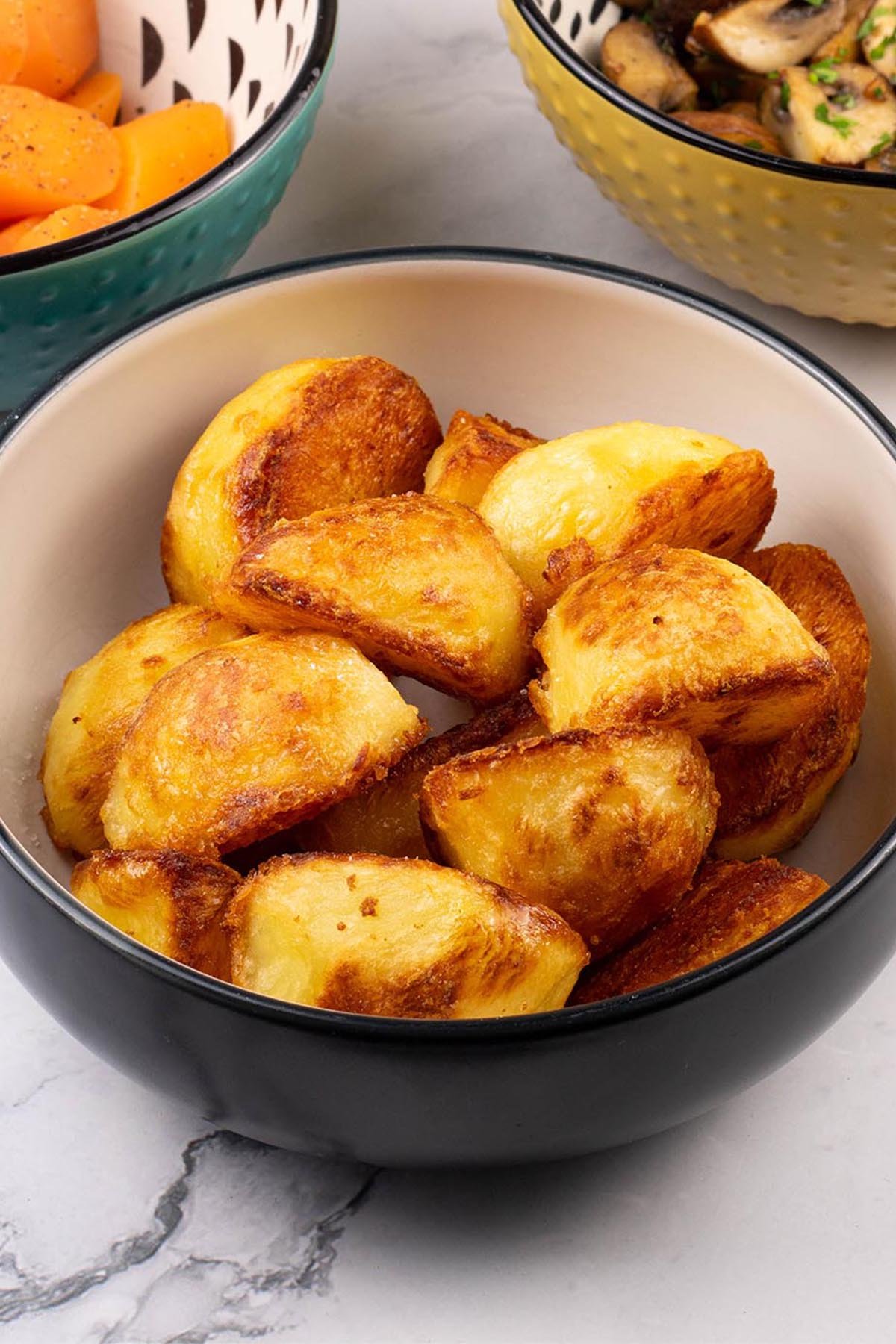 Roast potatoes in a black and cream serving bowl, with bowls of carrots and garlic mushrooms on the side.