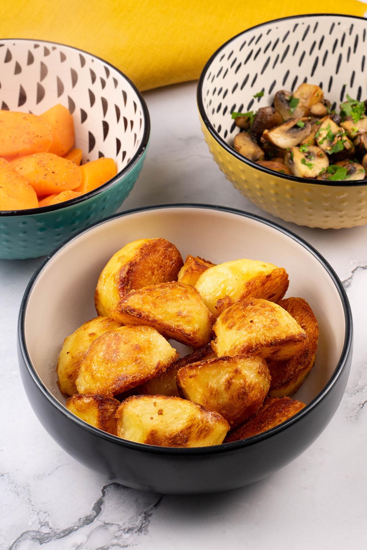 Roast potatoes in a black and cream serving bowl, with bowls of carrots and garlic mushrooms on the side.