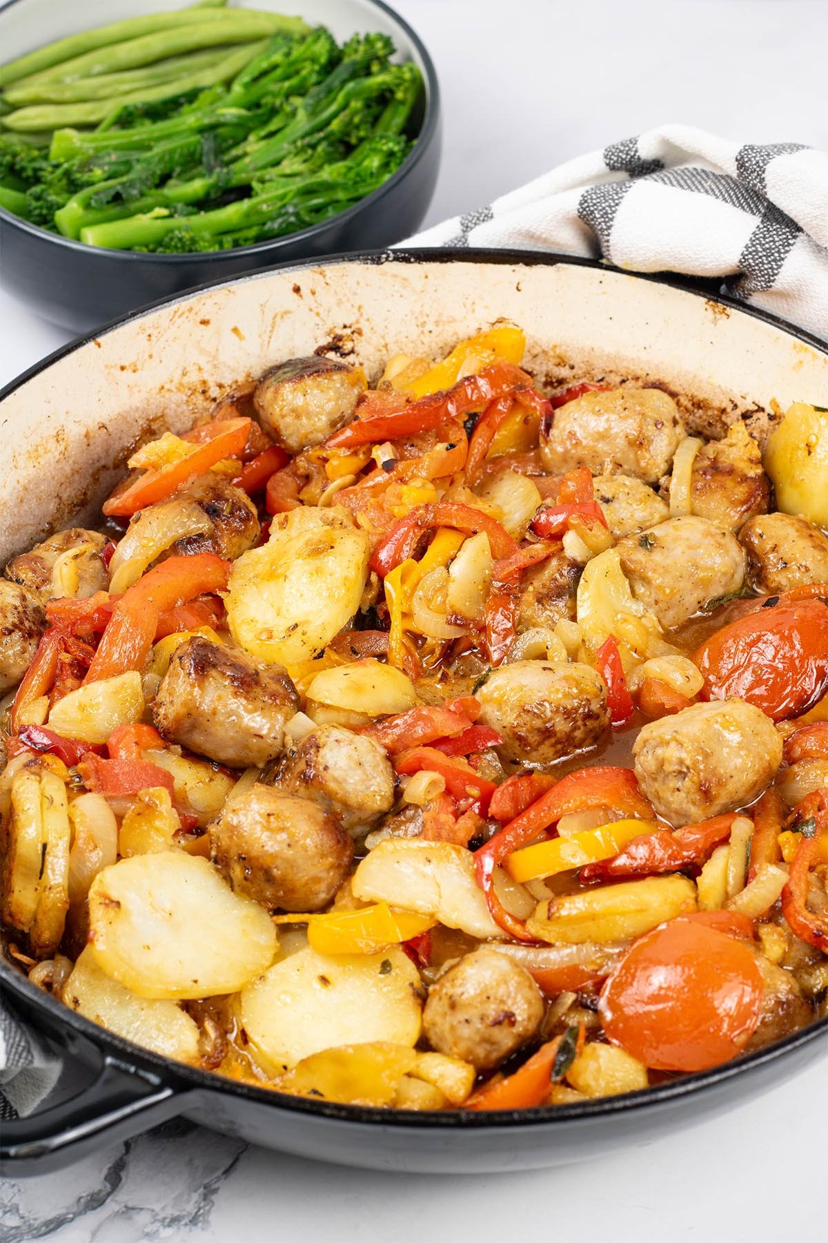 Salsiccia con patate e pomodori al forno in a large round casserole with a bowl of broccoli and green beans on the side.
