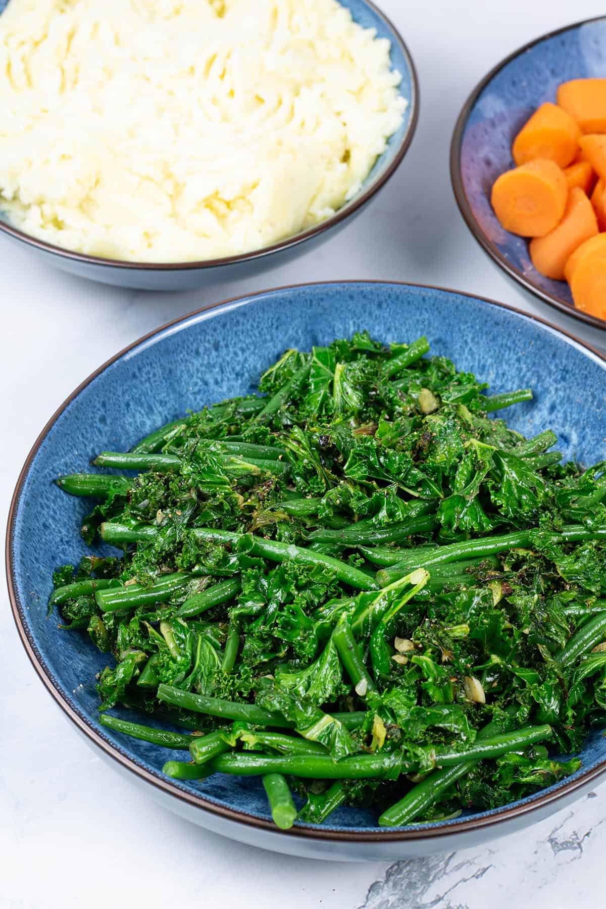 Sautéed kale and green beans in a blue patterned serving bowl, with bowls of mashed potatoes and quick cook carrots in the background.