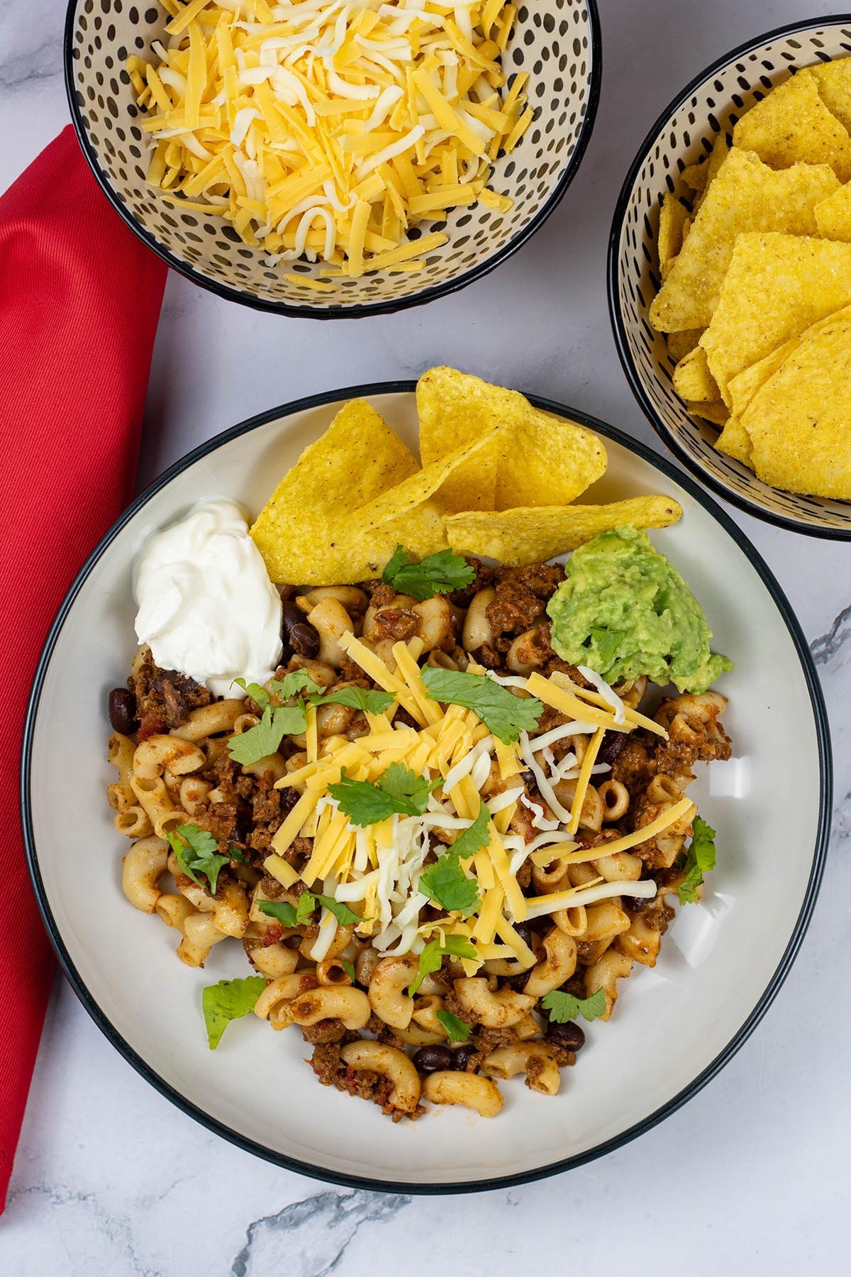 A portion of slow cooker cheesy chilli mac in a black and cream bowl with bowls of grated cheese and tortilla crisps on the side.