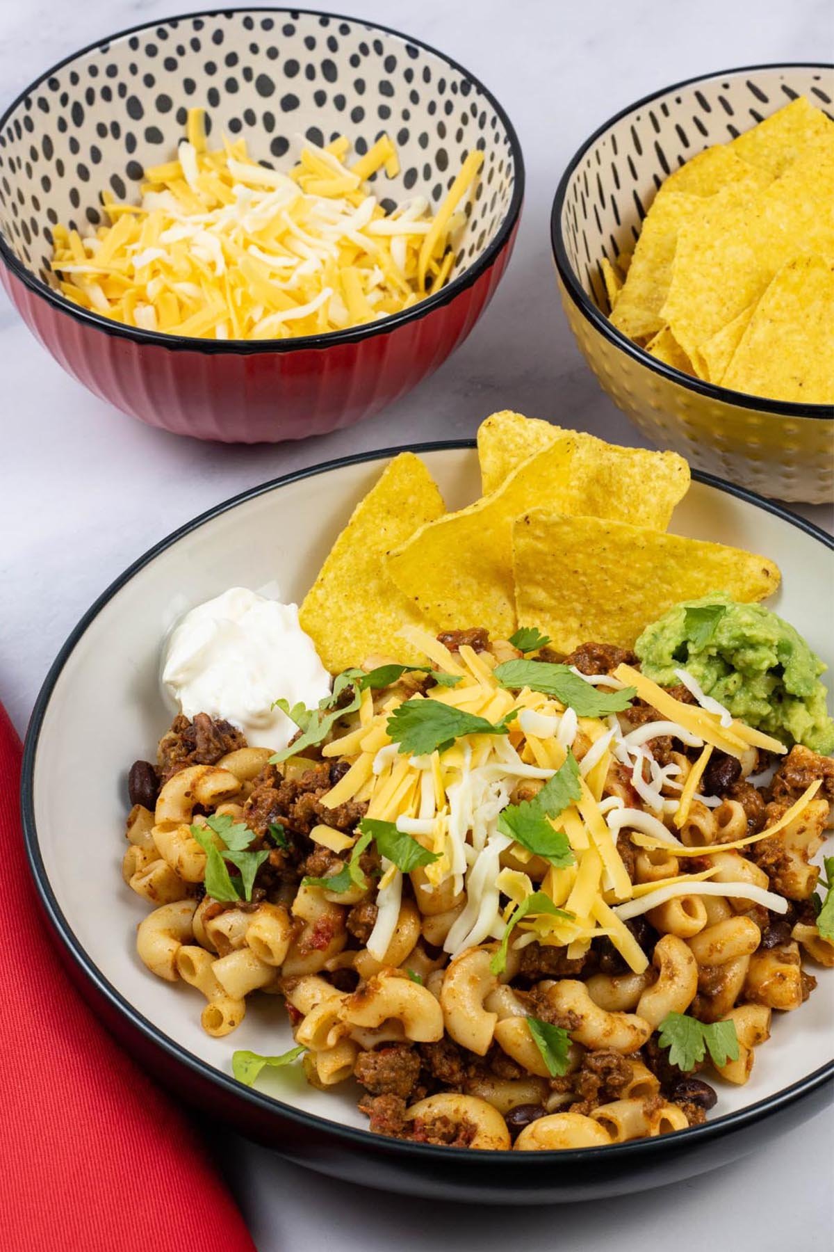 A portion of slow cooker cheesy chilli mac in a black and cream bowl with bowls of grated cheese and tortilla crisps on the side.