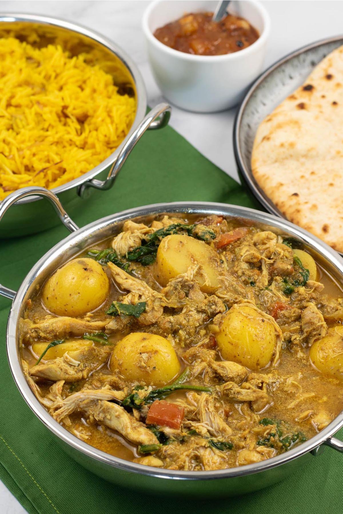 Slow cooker chicken and potato curry in a balti dish with golden pilau rice, chapatis and mango chutney in the background.