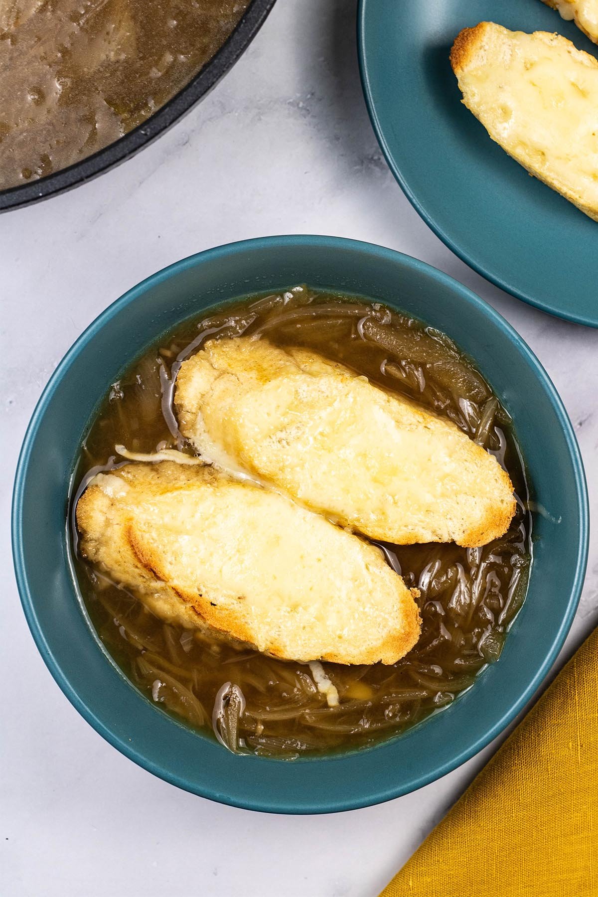 A portion of french onion soup in a teal bowl with 2 slices of baguette covered with melted cheese.