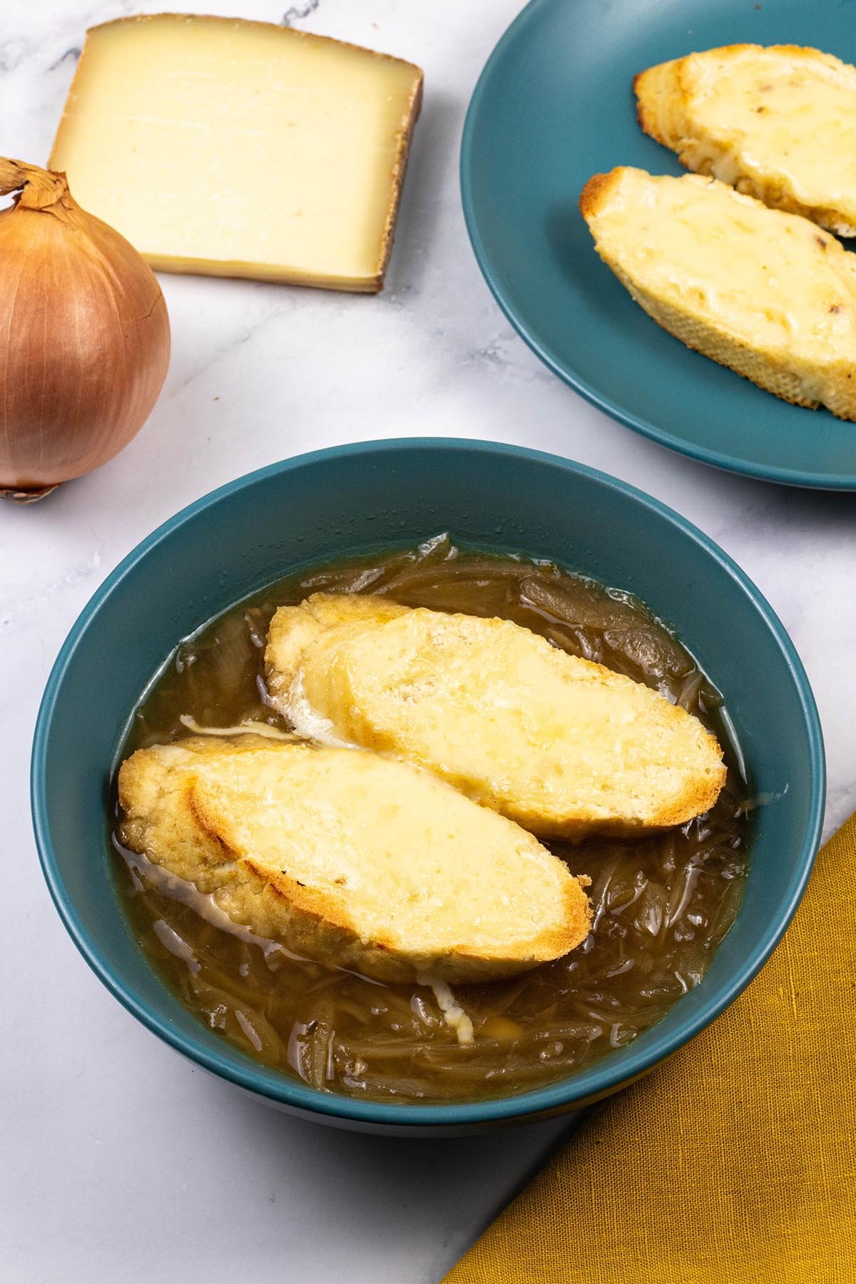 A portion of french onion soup in a teal bowl with 2 slices of baguette covered with melted cheese.