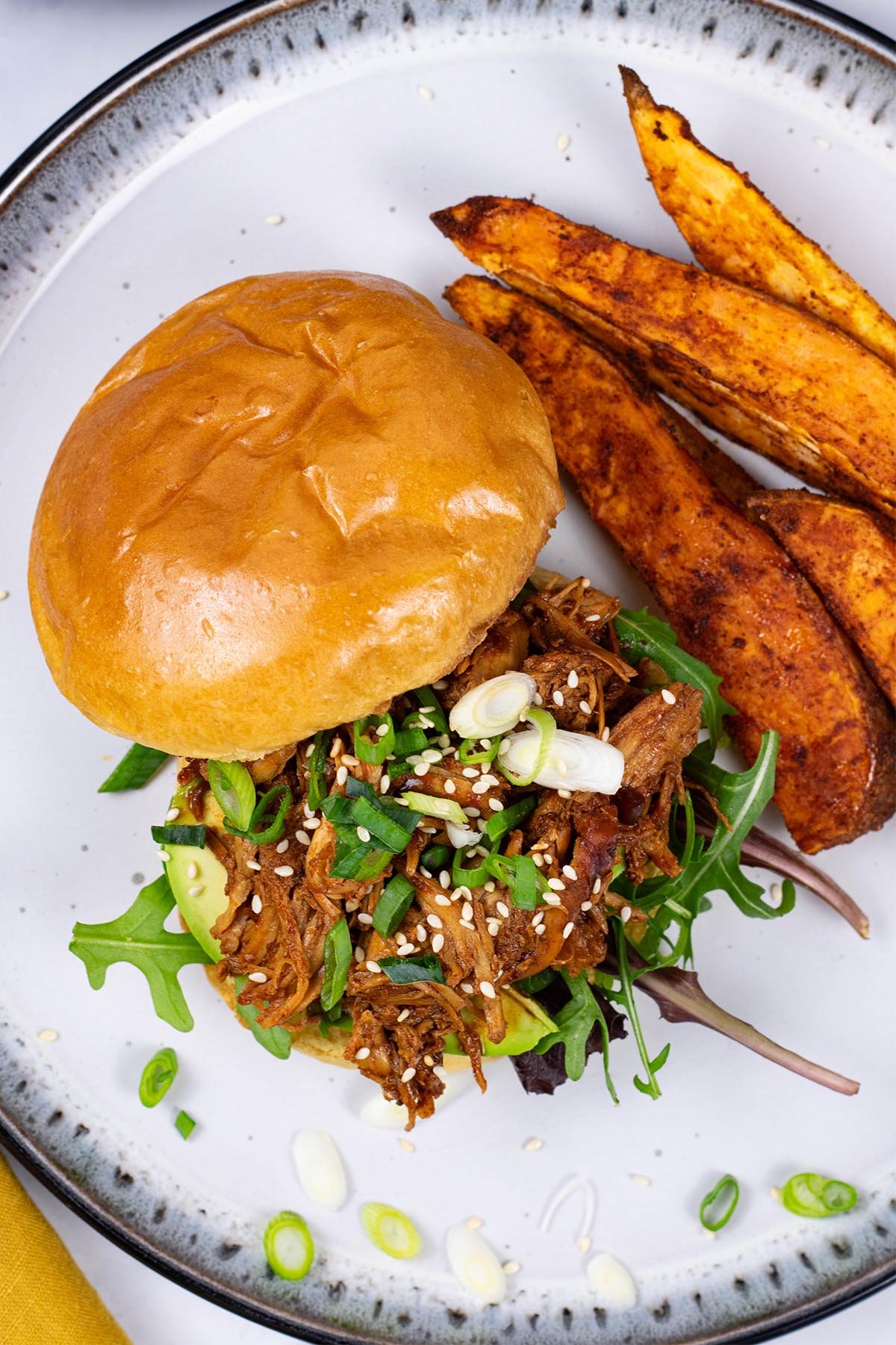 Easy slow cooker korean pulled chicken on a brioche bun with salad leaves, avocado slices, spring onions and toasted sesame seeds, and sweet potato wedges on the side.
