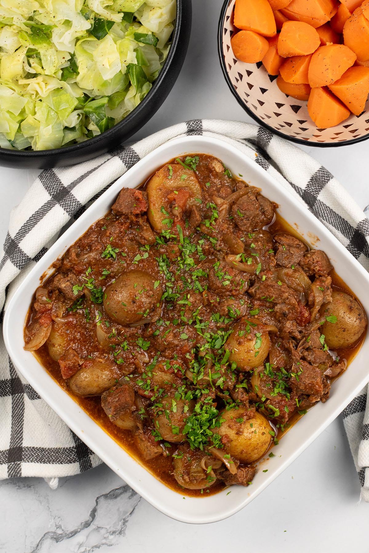 Slow cooker lamb stew with potatoes in a white square serving dish with bowls of buttered cabbage and quick cook carrots in the background.