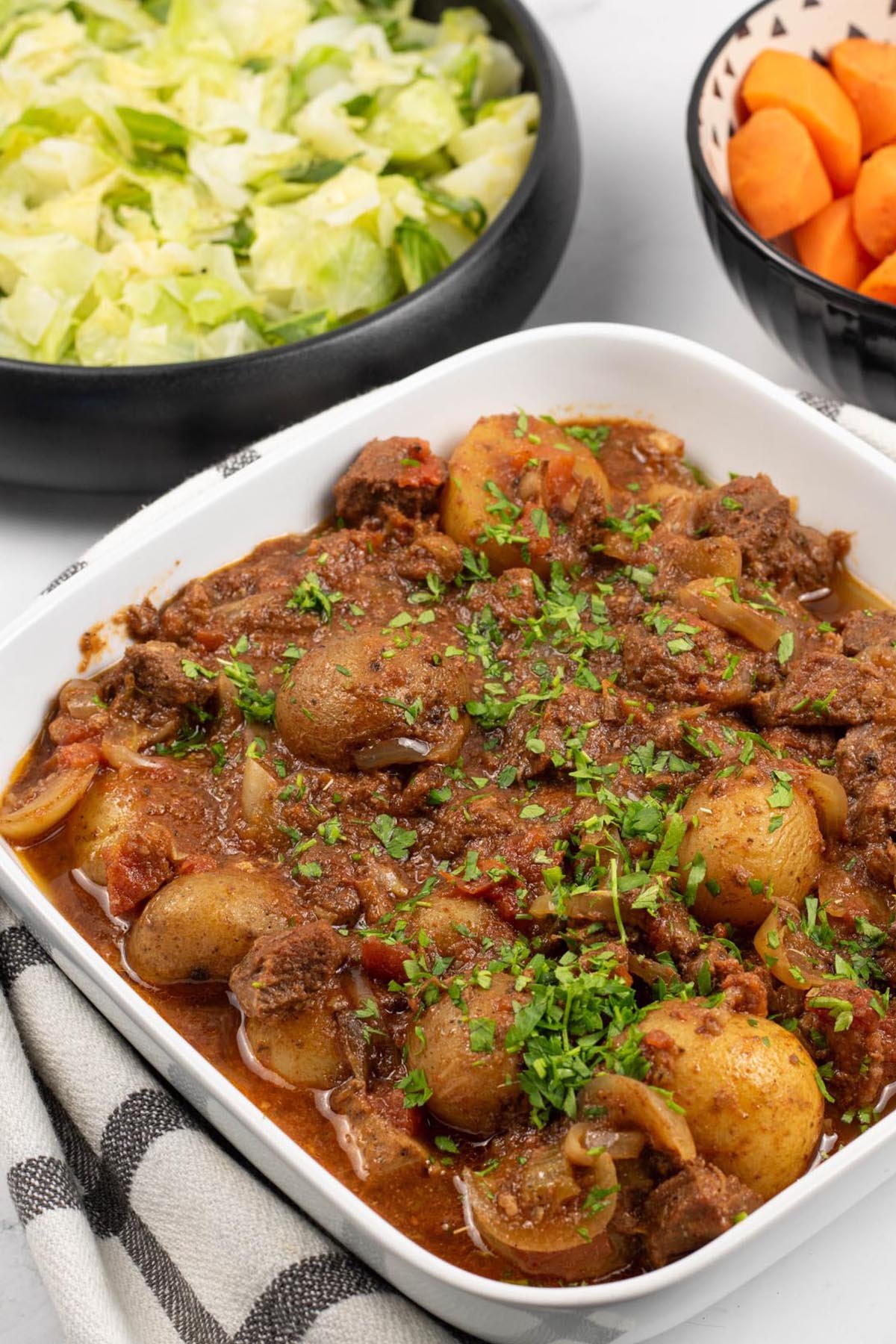 Slow cooker lamb stew with potatoes in a white square serving dish with bowls of buttered cabbage and quick cook carrots in the background.