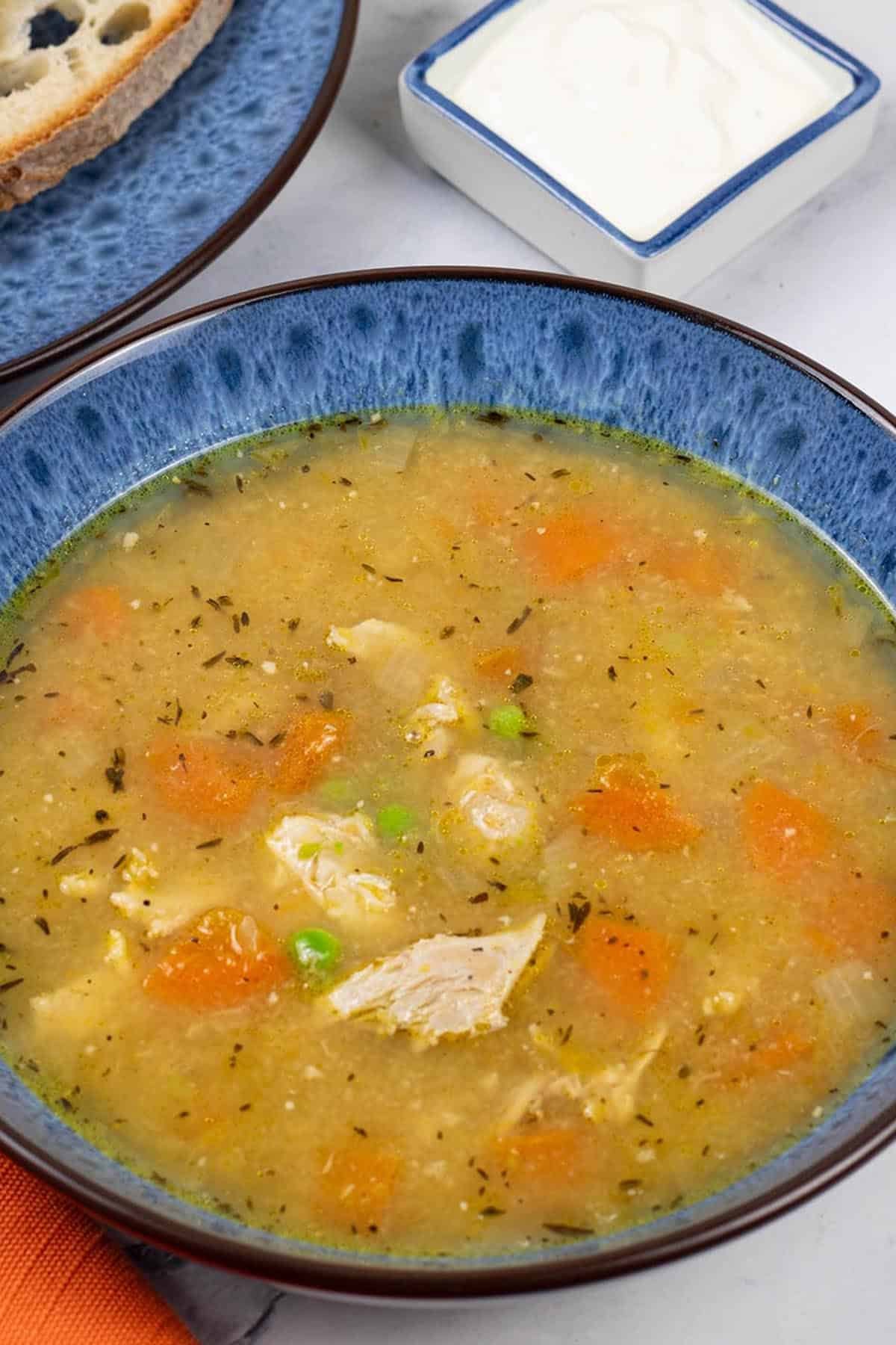 A portion of slow cooker roast chicken soup in a blue patterned bowl with a plate of sliced Portuguese water bread and dish of garlic and lemon yogurt on the side.